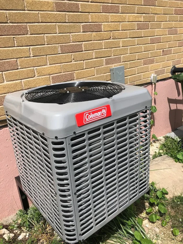 An outdoor air conditioning unit by Coleman next to a brick wall and pink siding, with some plants and grass nearby.