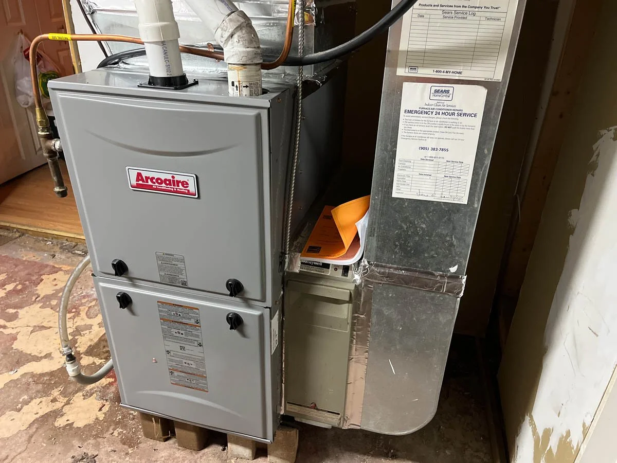 A silver Arcoaire heating, ventilation, and air conditioning unit installed on a wooden pallet in a utility room, with several pipes and ducts connected to it, and various service and emergency contact labels posted nearby.