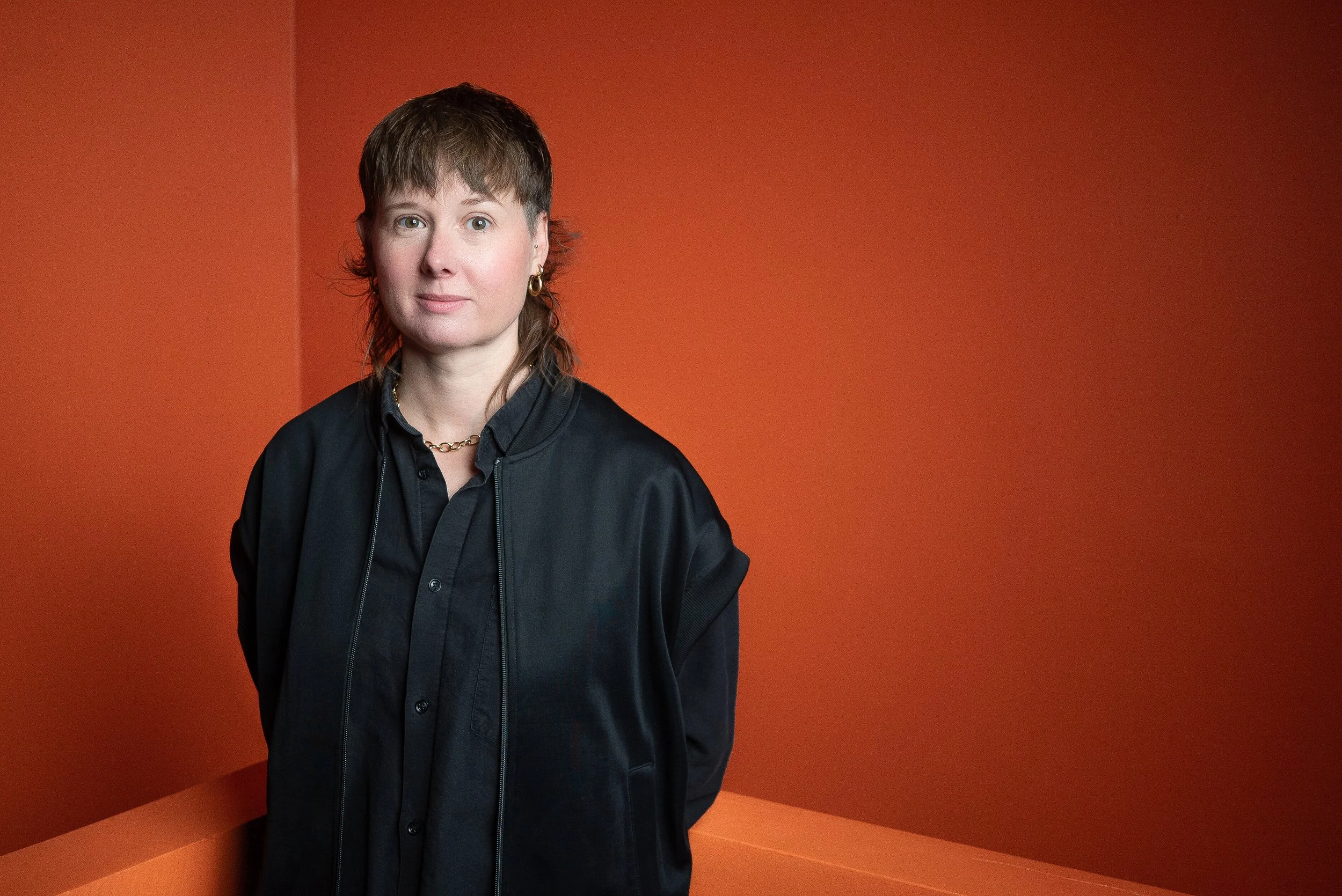 A woman with short brown hair wearing a black shirt and jacket, gold hoop earrings, and a gold chain necklace standing in front of an orange background with geometric shapes.