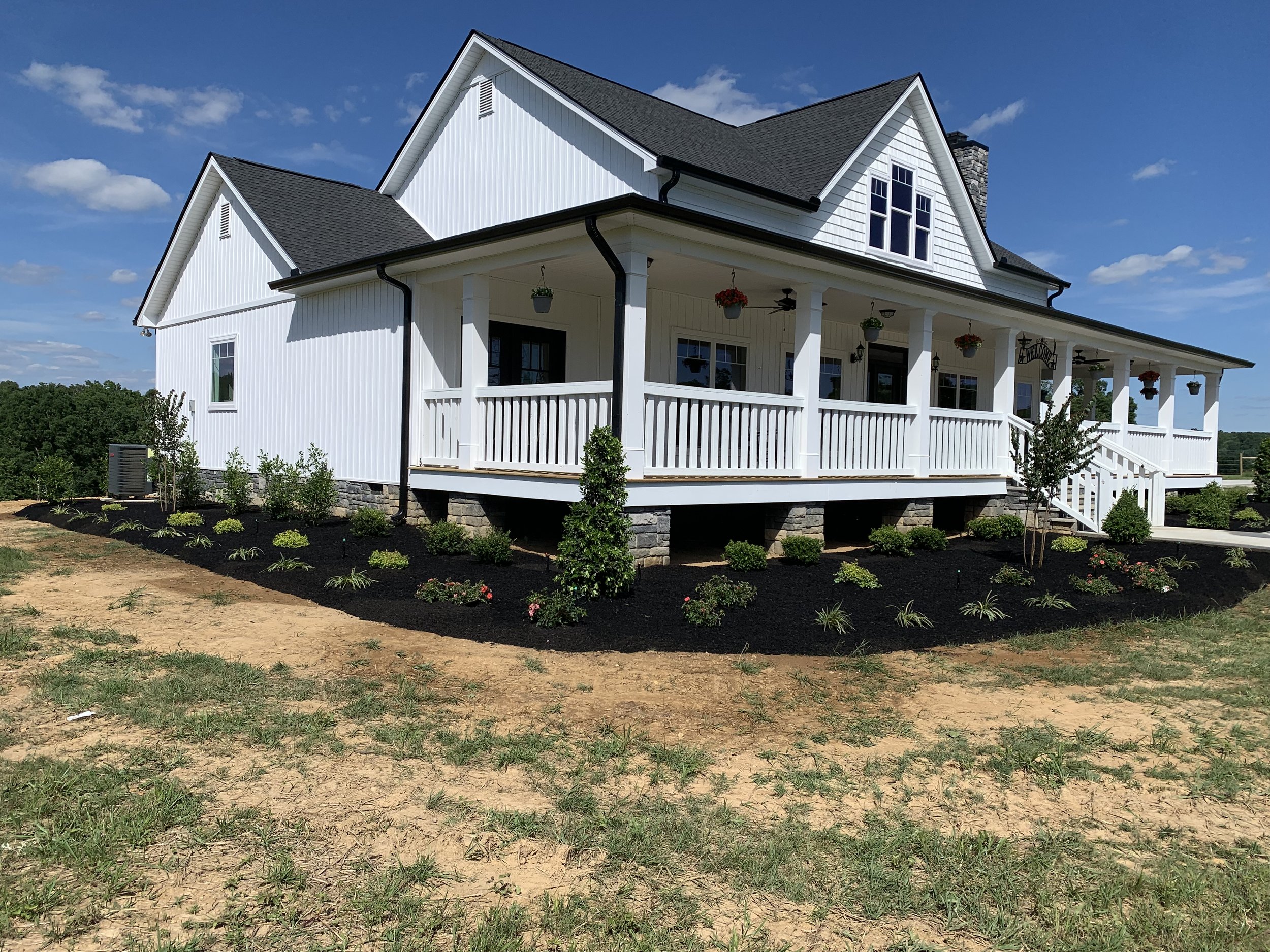 White two-story house with a covered front porch, black roof, and flower pots hanging from the porch ceiling, surrounded by a newly landscaped yard with small plants and shrubs.
