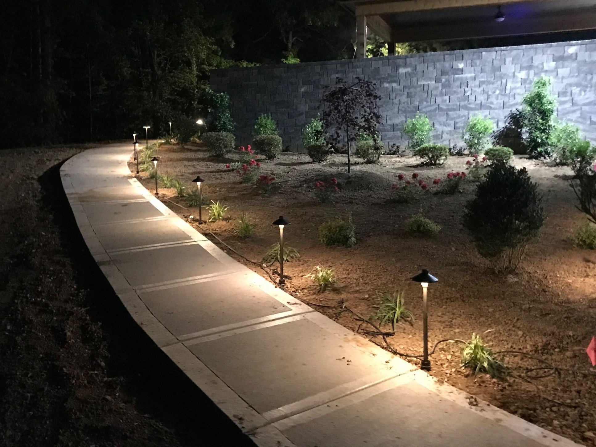 Nighttime view of a curved sidewalk with small landscape lights, surrounded by a landscaped garden with various bushes and a stone wall in the background.