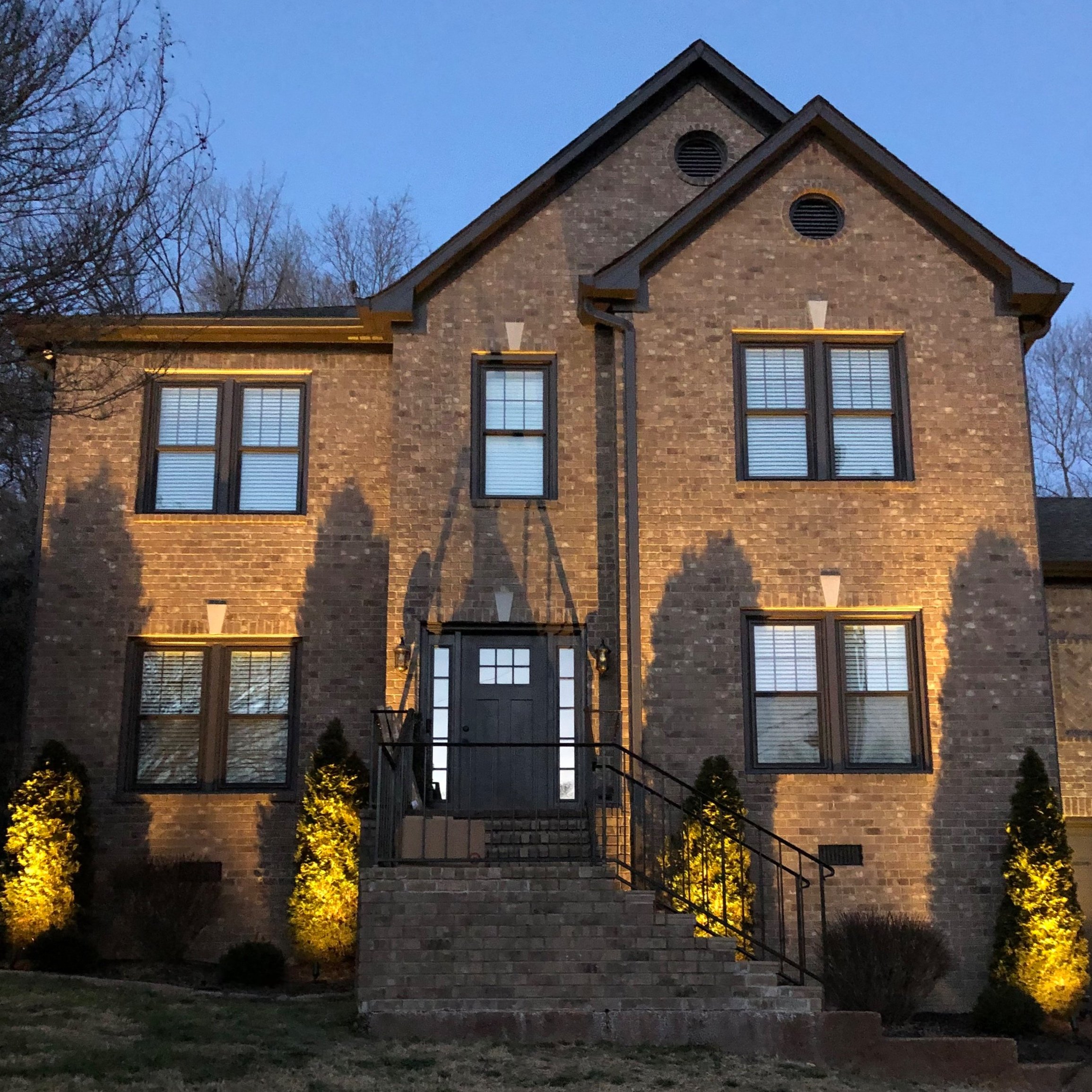 A two-story brick house at dusk with illuminated landscaping and windows.
