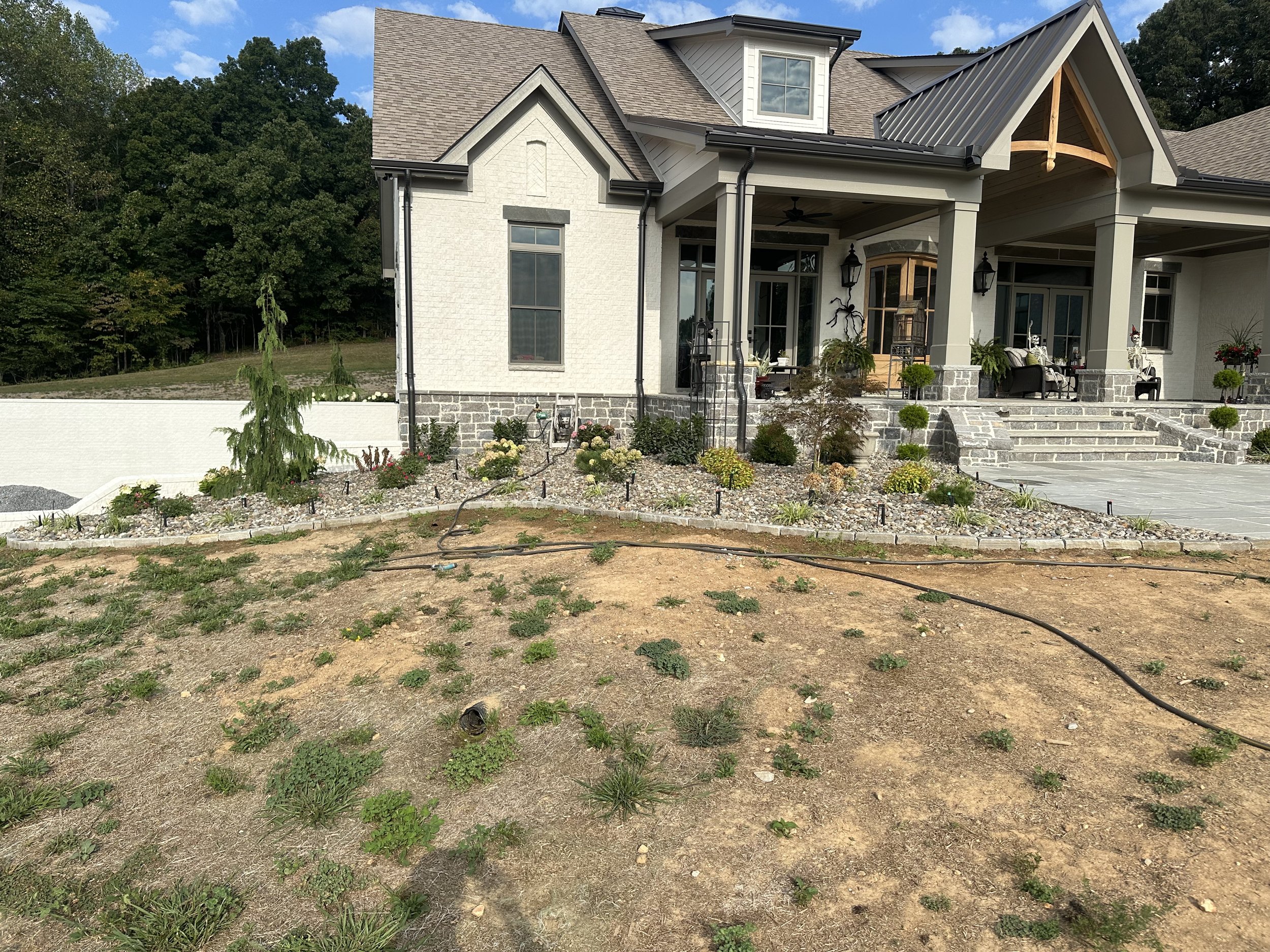 A modern house with a landscaped front yard, featuring a mix of stone and garden beds, with some plants and grass patches, and a porch with outdoor furniture and lights.