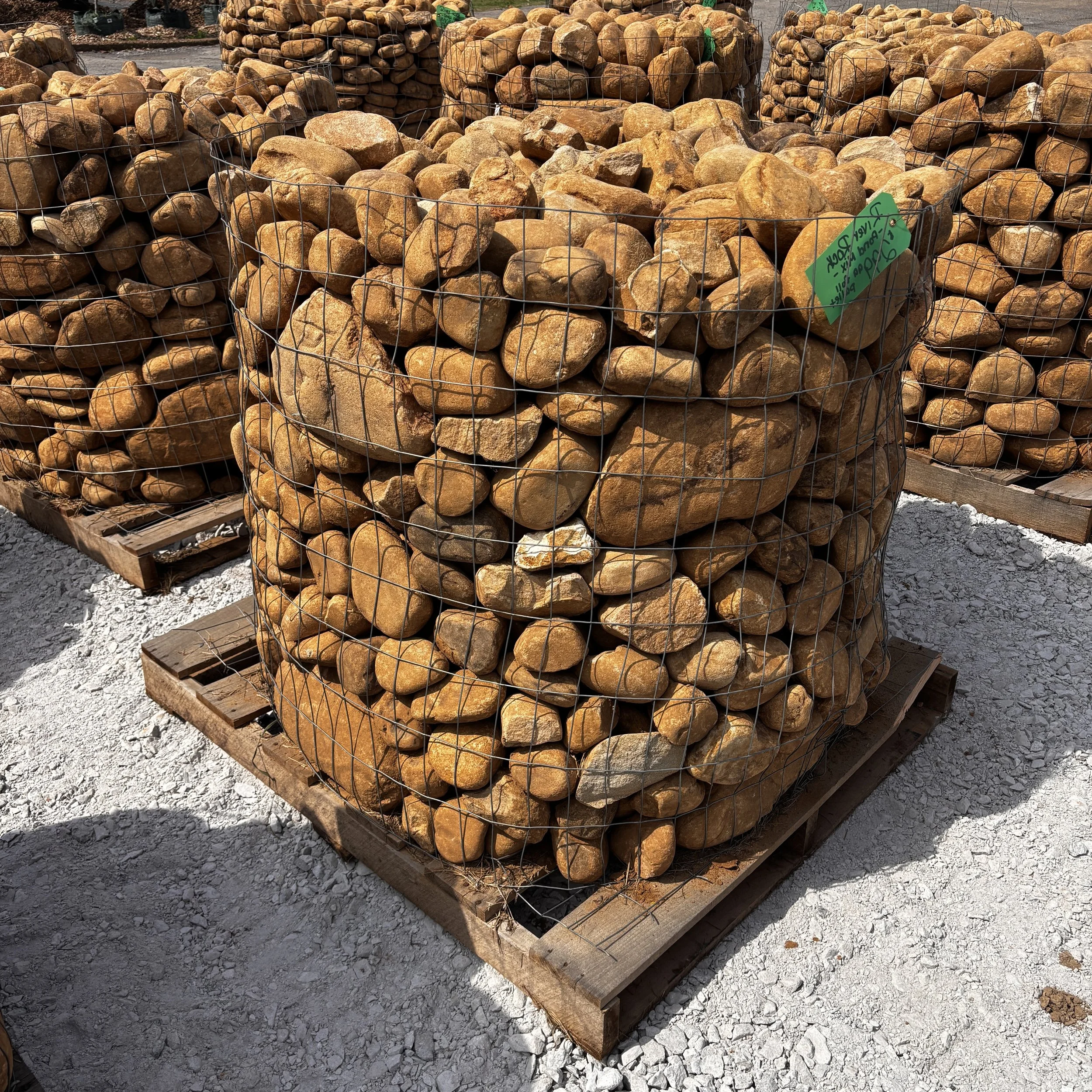 Stacks of large, rounded tan and brown stones arranged in wire cages on wooden pallets at an outdoor hardware store or landscape supply yard.