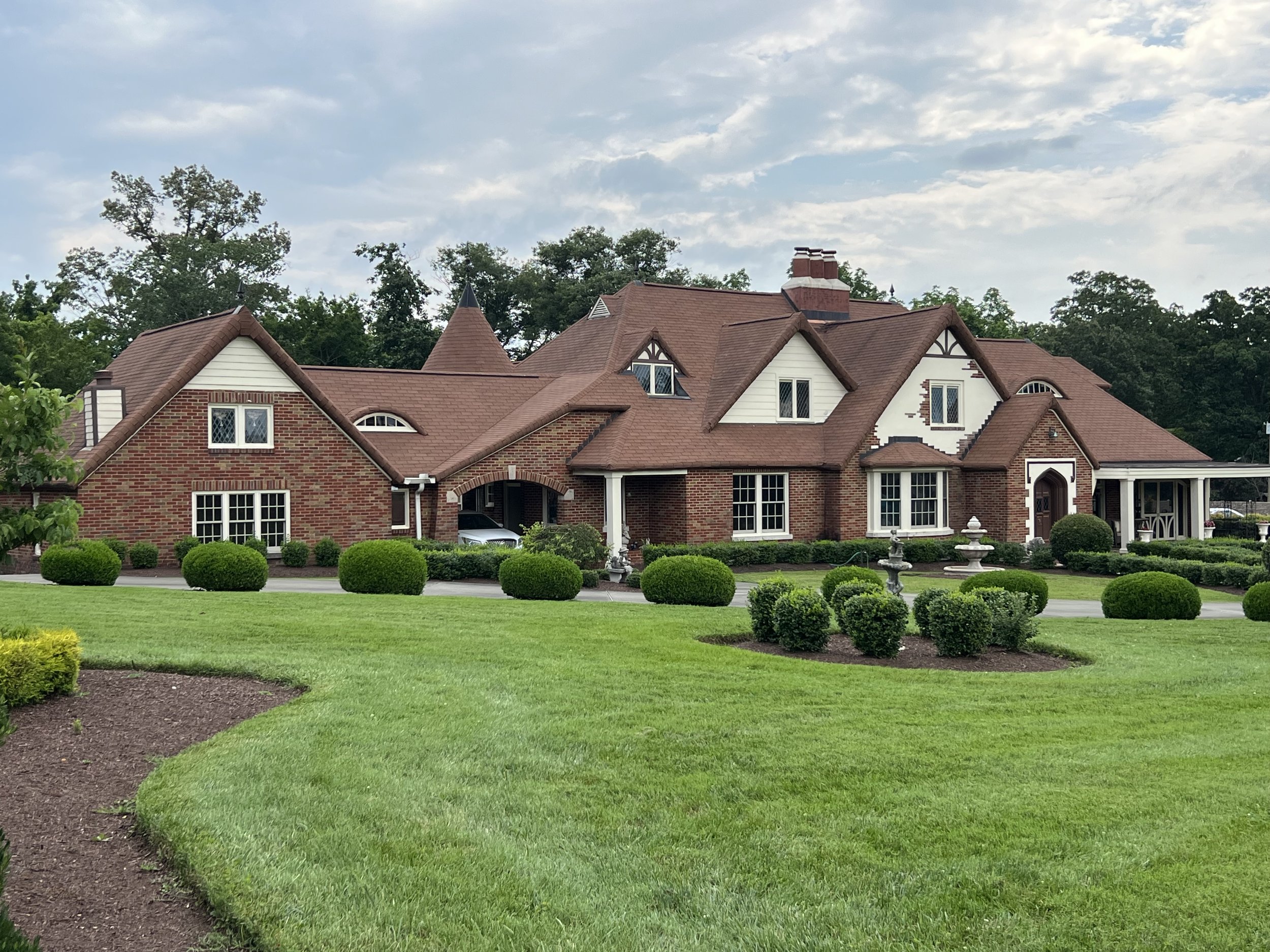 Large mansion with red brick walls, multiple gabled roofs with brown shingles, surrounded by neatly landscaped lawn and bushes, and a fountain in front.