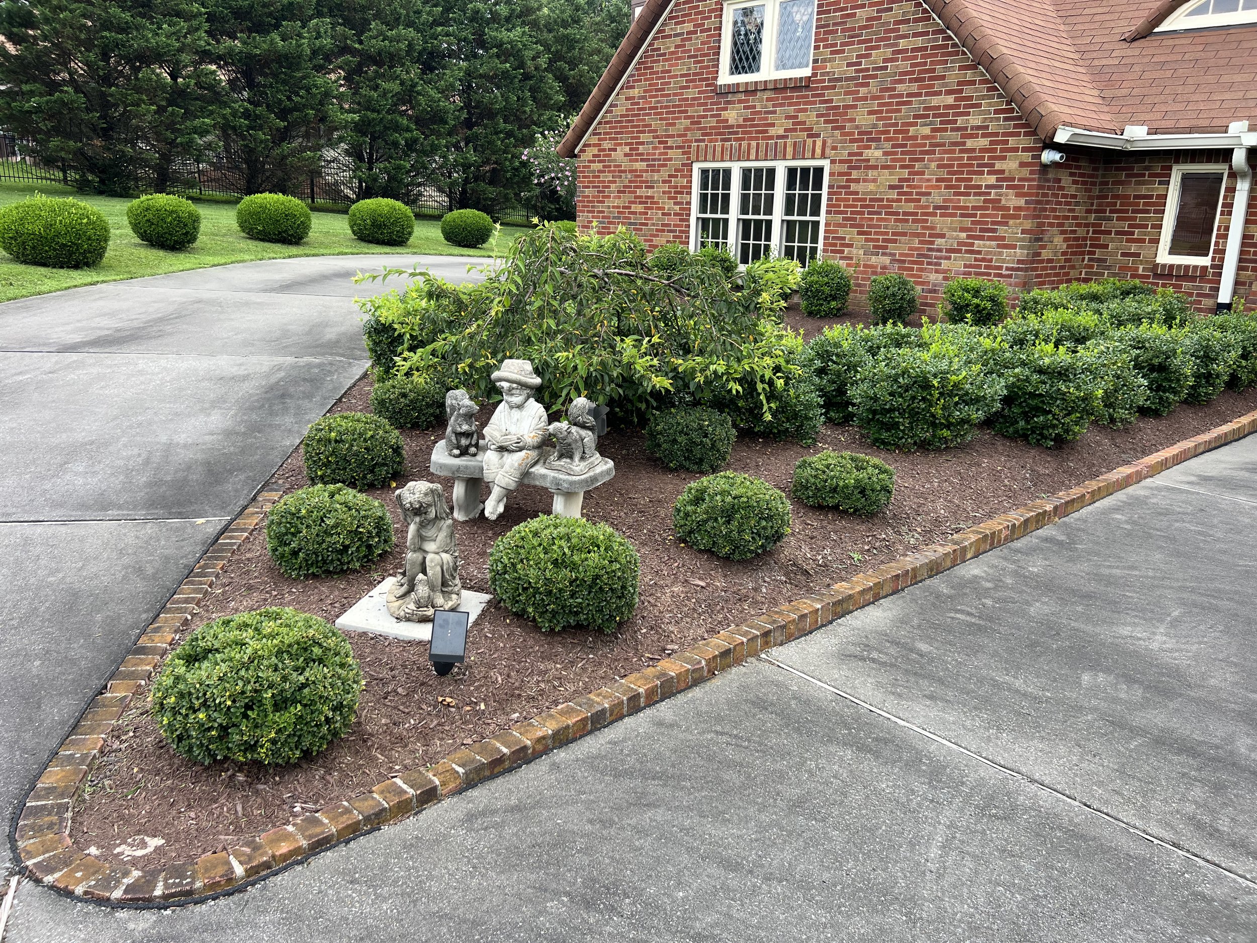 Front yard garden with neatly trimmed bushes, stone statues including a person with children, and a brick house with white window frames.