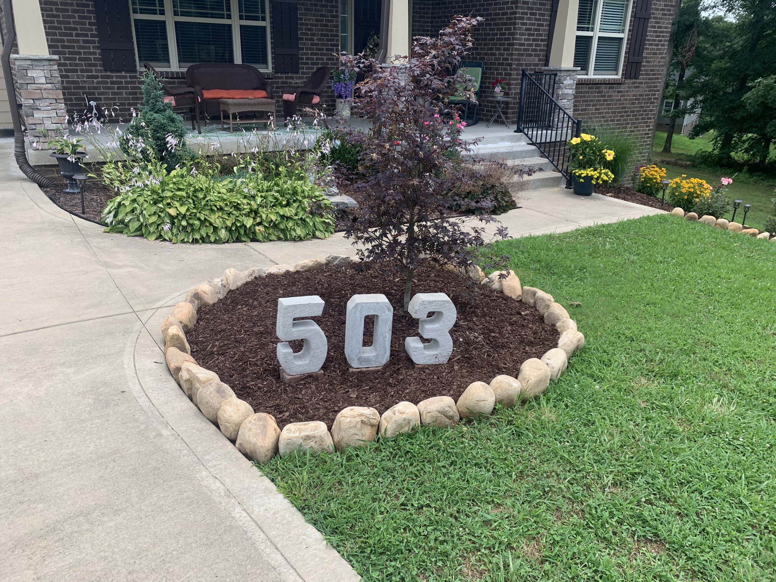 A house front yard with a flower bed bordered by stones, displaying house number 503 in large concrete numbers, a small tree, and a manicured lawn.