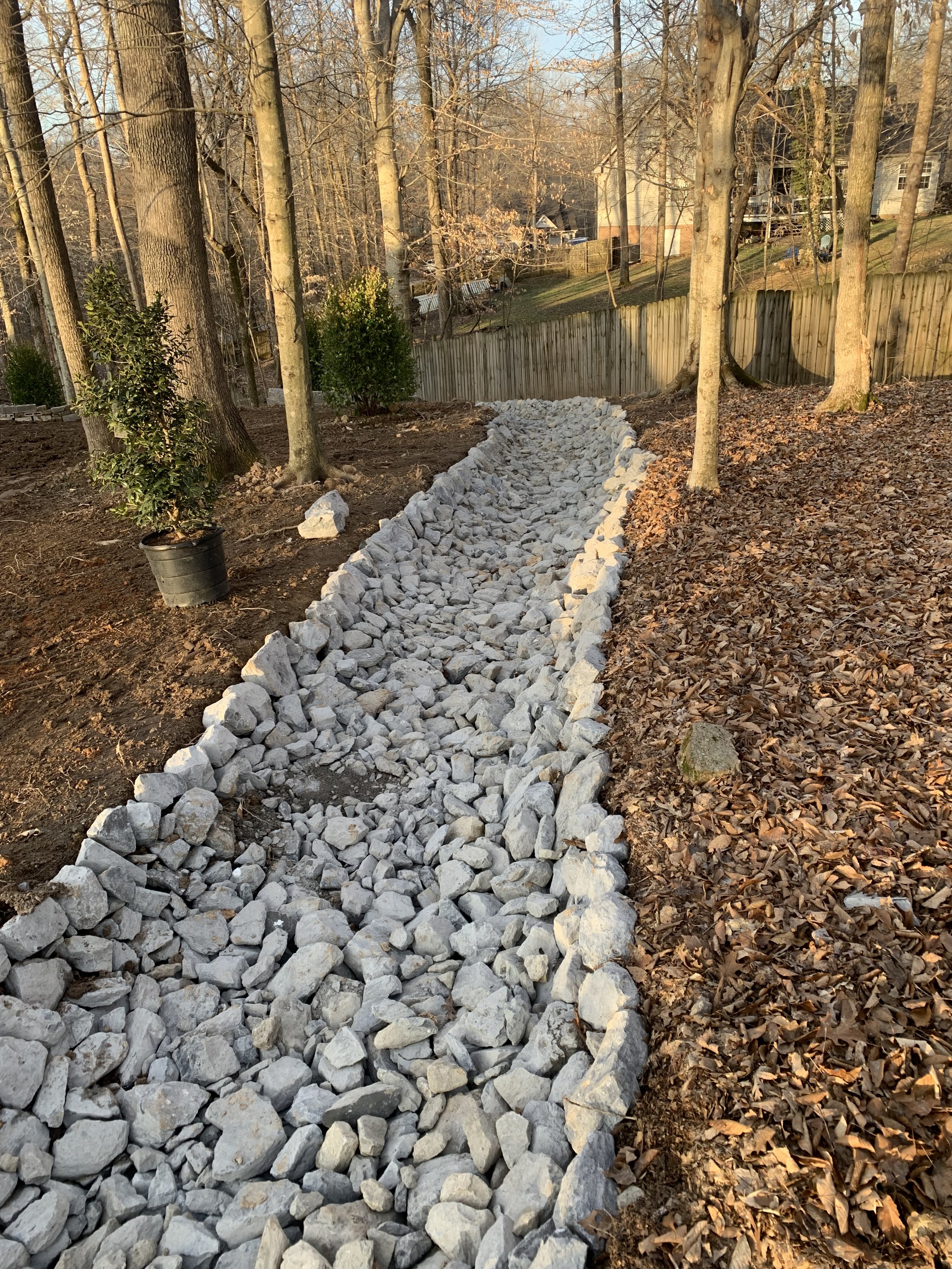 A gravel walkway under construction in a backyard, bordered by rocks and trees, with a wooden fence in the background.