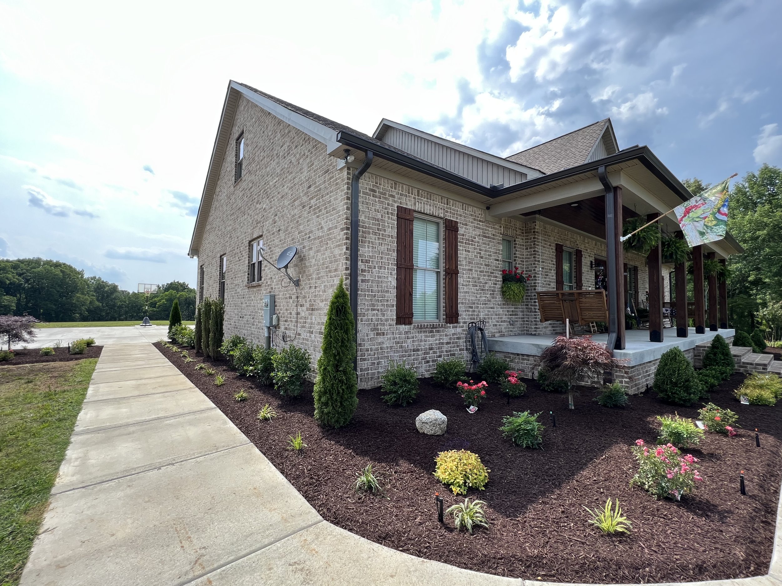 Front exterior of a brick house with a landscaped garden, porch, and flag, on a partly cloudy day.