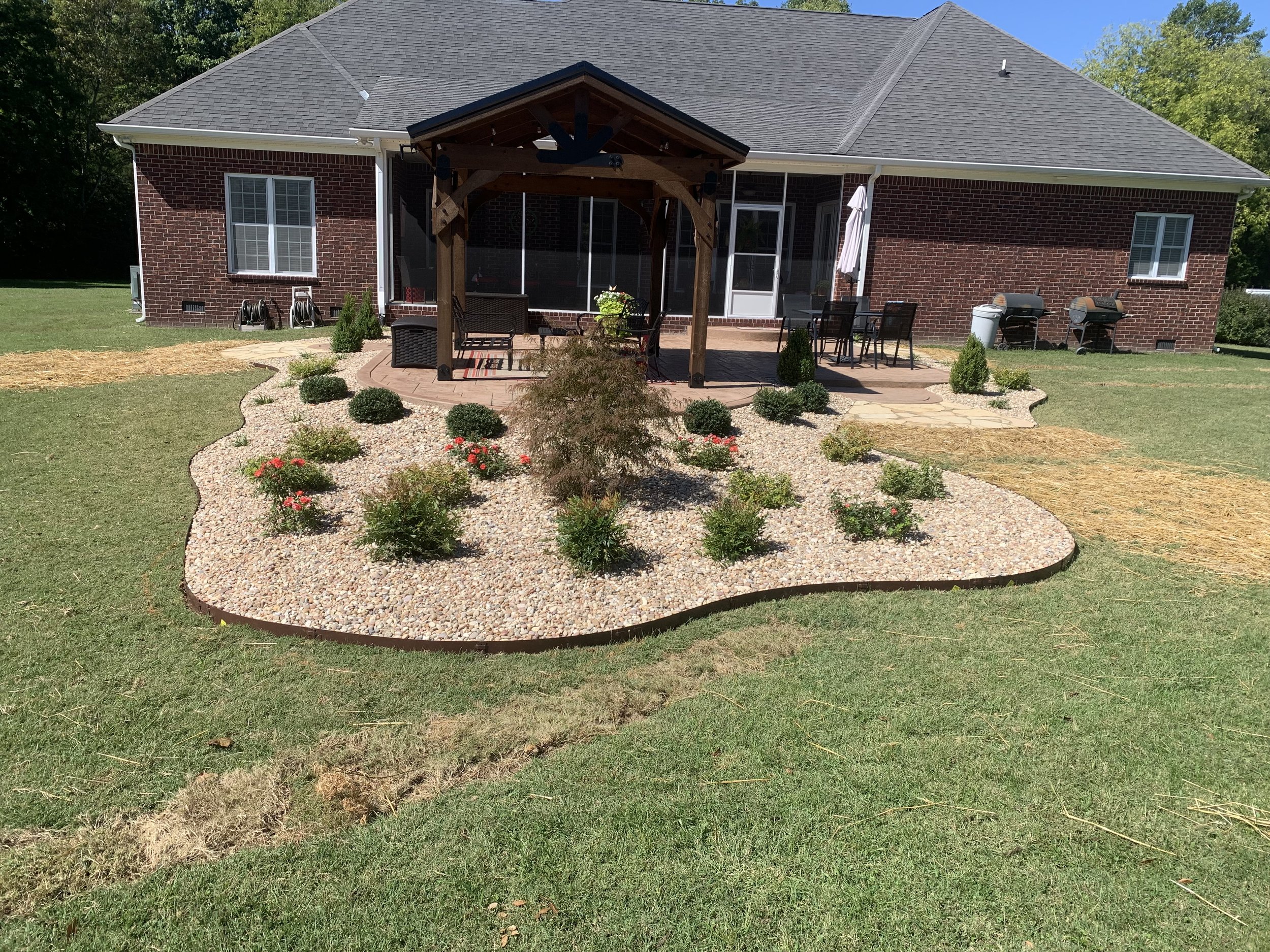Backyard with a landscaped flower bed, small bushes, and flowers, surrounded by a curved border, with a brick house and a covered patio area with outdoor furniture in the background.