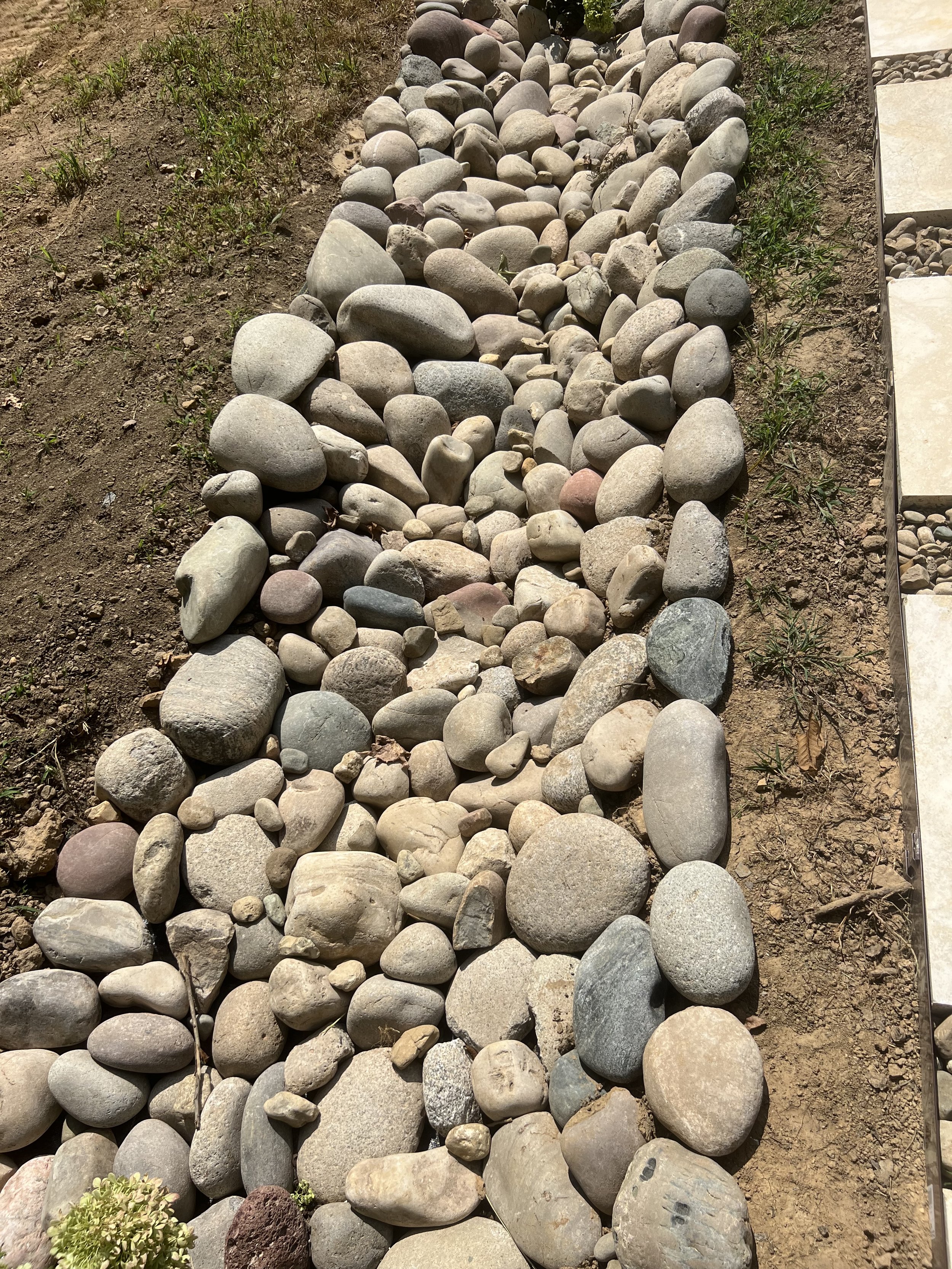 A dry riverbed made of various-sized smooth stones bordered by dirt and some grass, with concrete tiles visible on the right side.