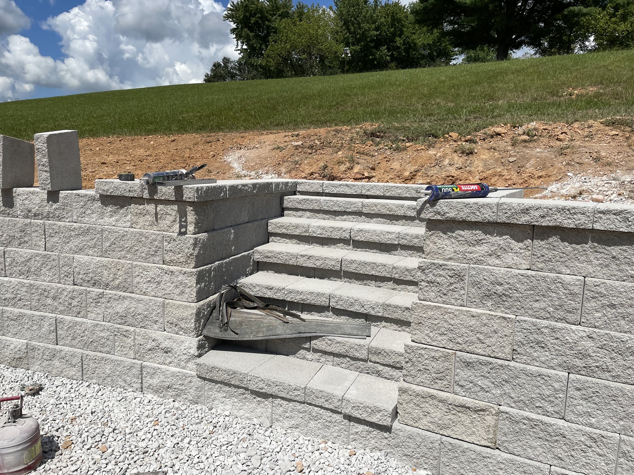 Construction site with gray stone retaining wall and stairs, tools on top, and a grassy hill with trees in the background.