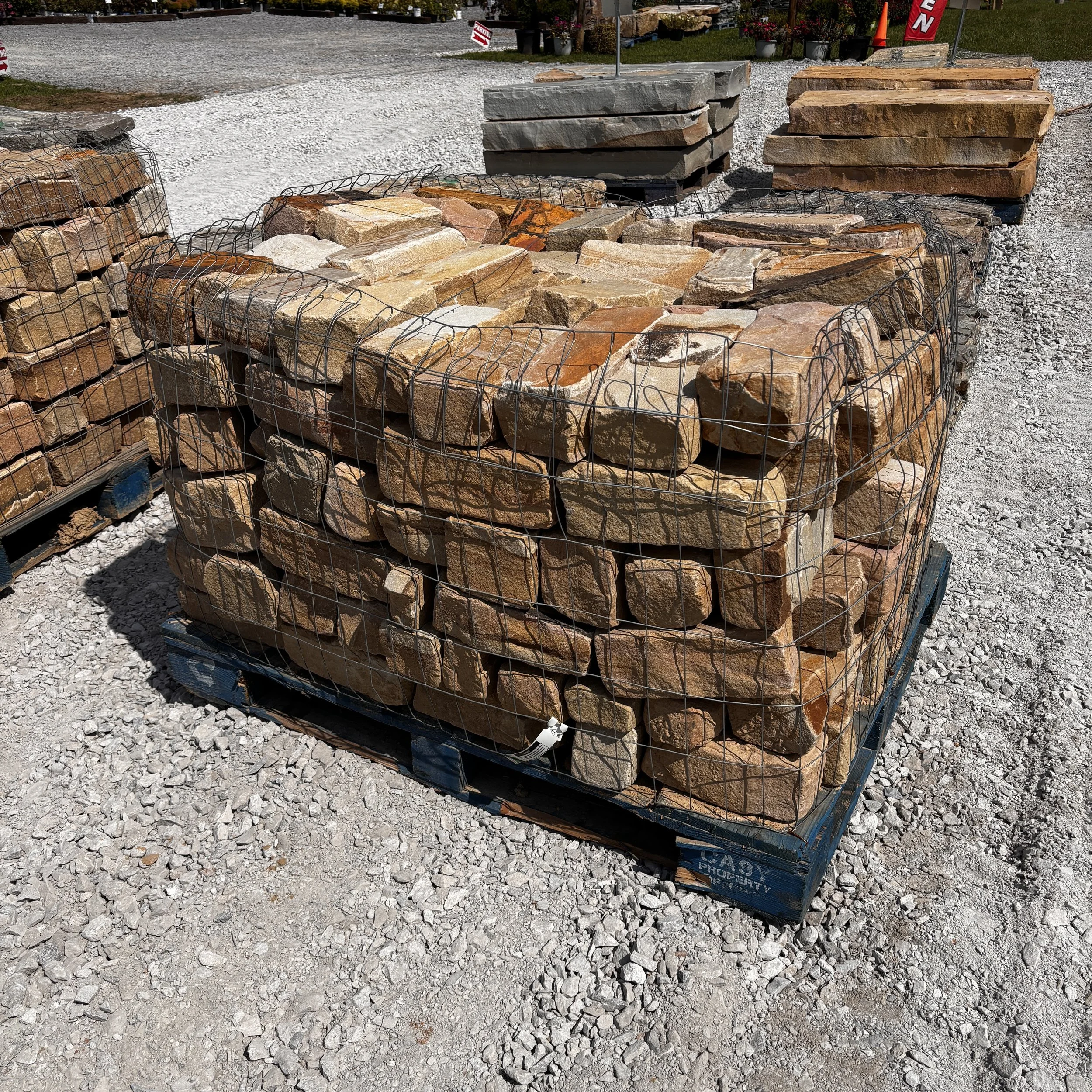 Stacks of different types of large natural stone bricks on pallets at a garden center or hardware store with gravel and dirt in the foreground and plants in the background.