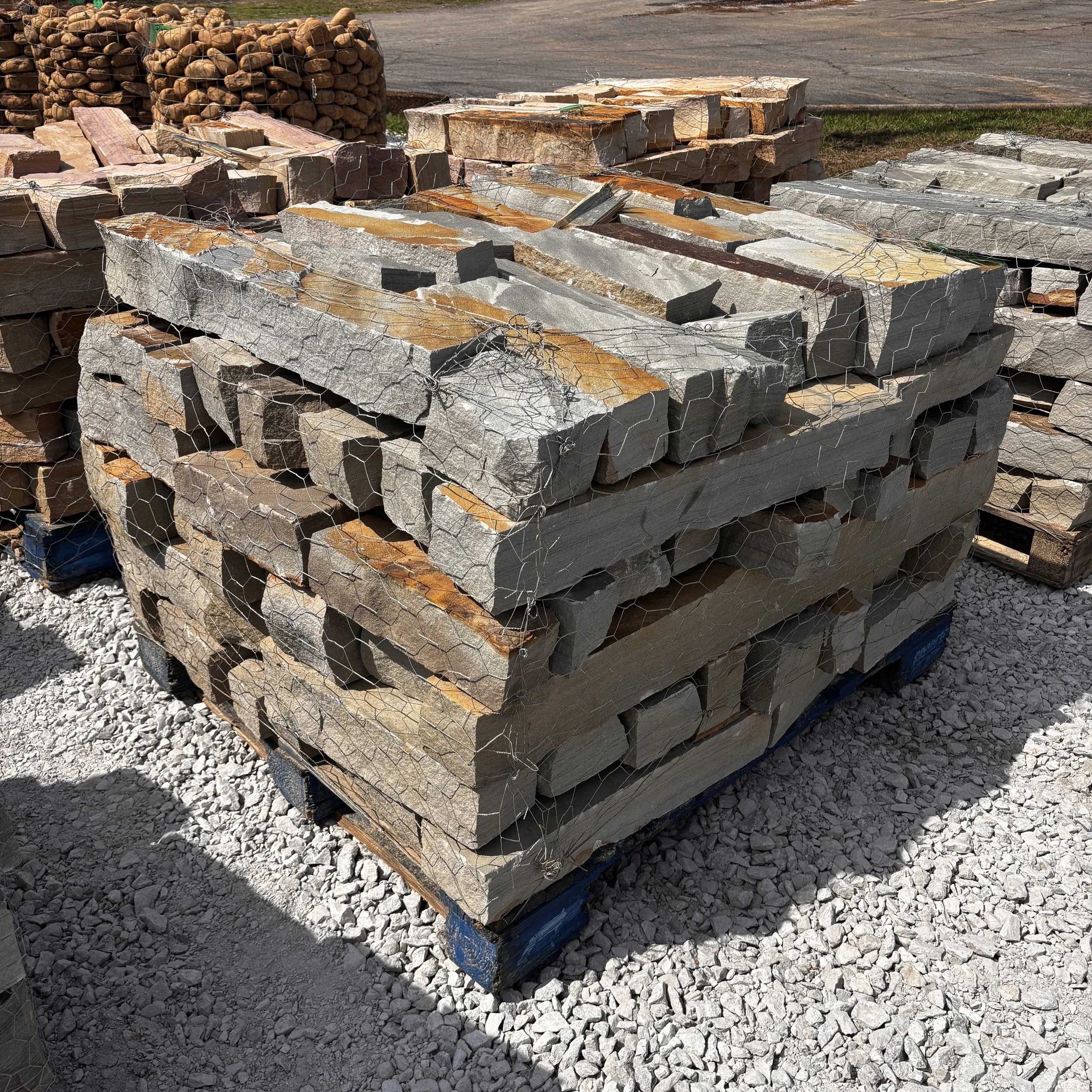 Piles of large natural stone slabs and stacks of rounded stones behind wire fencing at an outdoor construction or landscaping site.