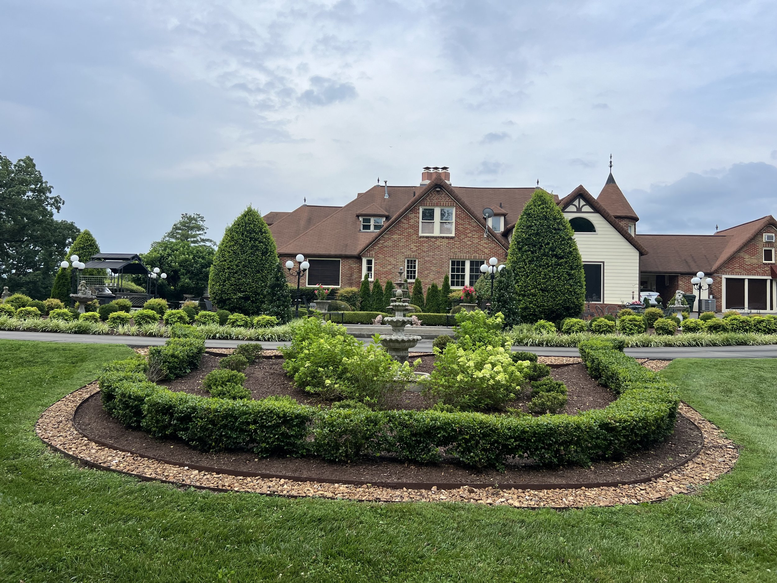 Large, well-maintained lawn with neatly landscaped flower bed featuring trimmed bushes and trees in front of a large brick house with brown roof and decorative windows.