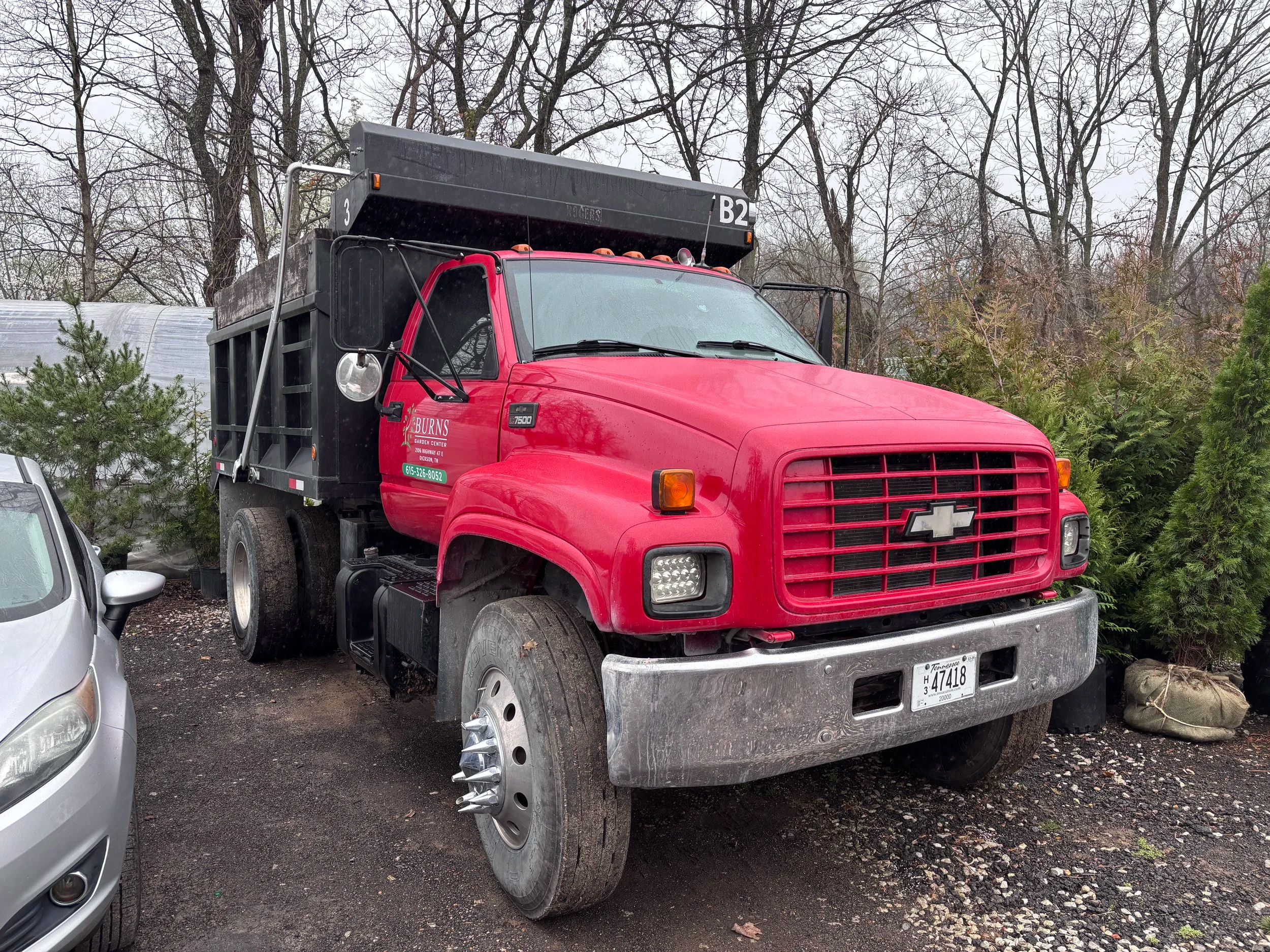 Red utility truck parked in a lot next to a silver car, with trees and some shrubbery in the background.