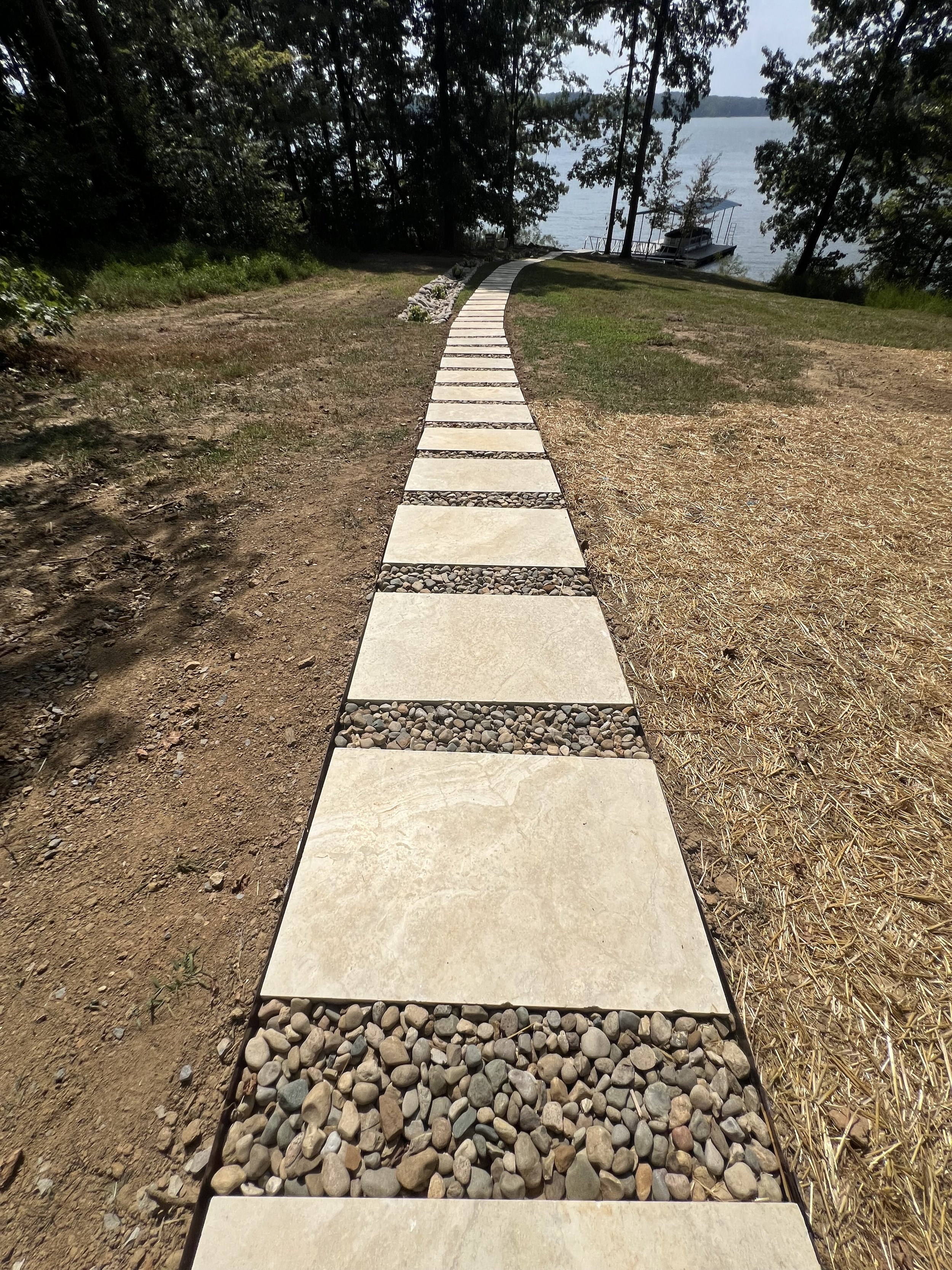 Concrete walkway with stone accents leading to a lake with trees and a boat in the background.