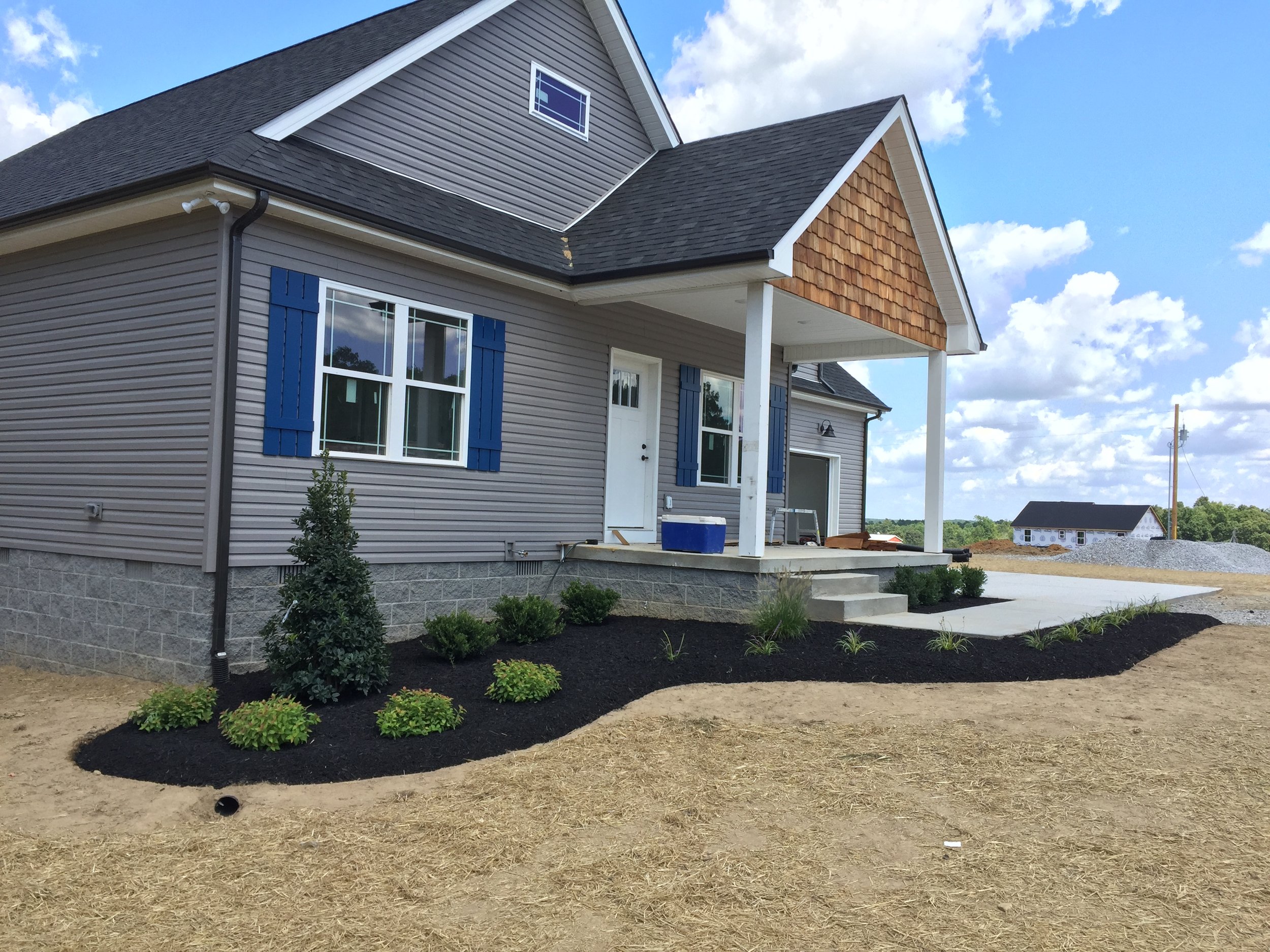 Newly constructed two-story house with gray siding, blue shutters, and a front porch with steps leading up to the entrance. There are small landscaped bushes and plants in front of the house, with some areas of bare dirt and a partly cloudy sky above