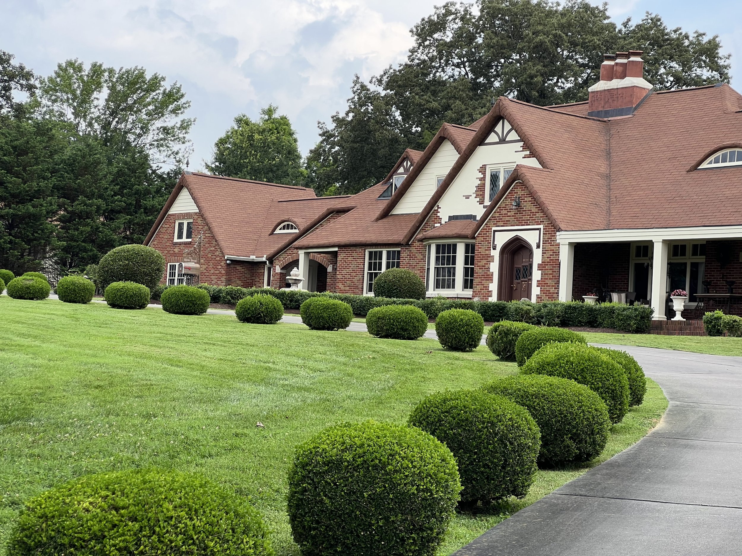 Large brick house with a red roof, surrounded by a green lawn and neatly trimmed bushes, with a curved driveway leading to the entrance.