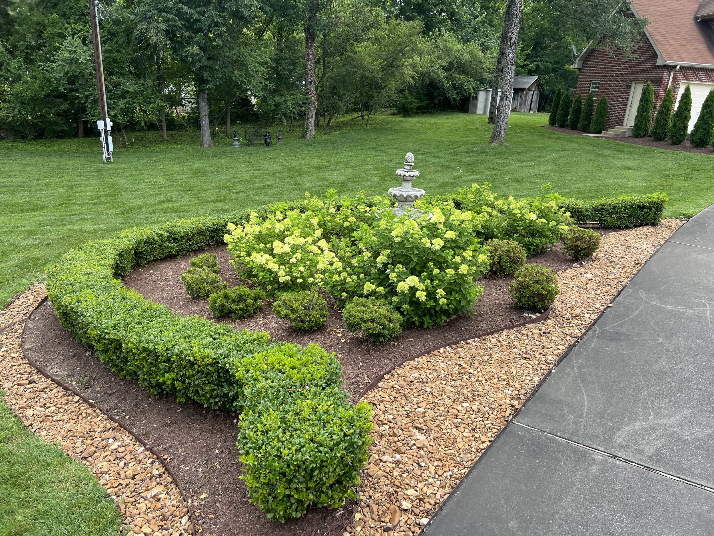 Landscaped garden with a white decorative fountain, green shrubs, hydrangea flowers, gravel, grass, trees, and a house in the background.