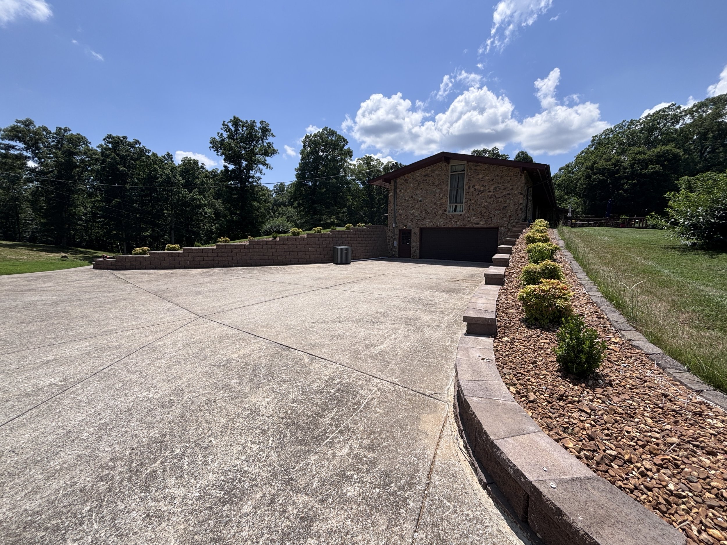 A residential driveway with a stone house in the background, a flower bed with small bushes on the right, and a clear blue sky with some white clouds overhead.