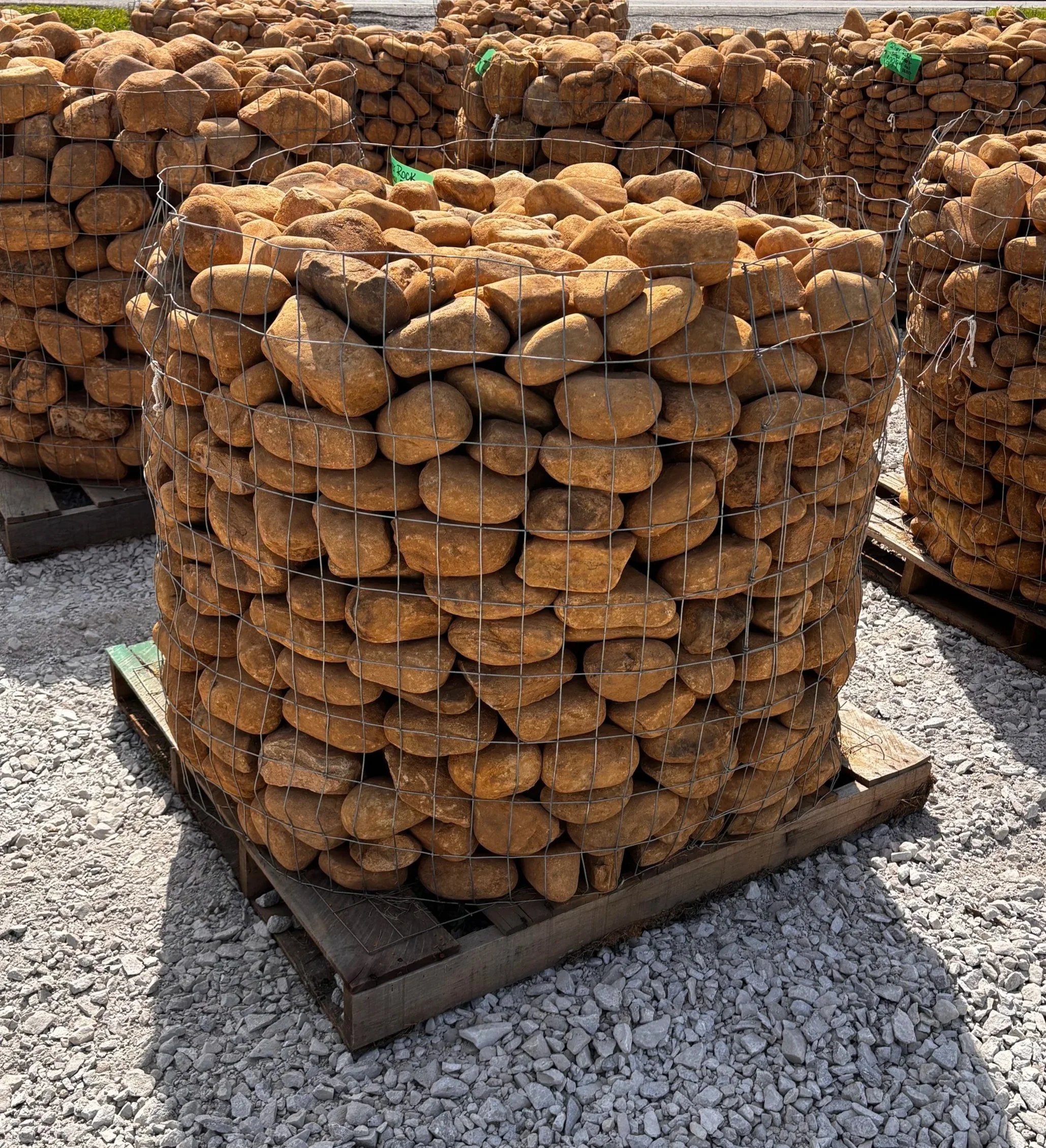 Large stacks of imported river rocks and pebbles held in wire cages, placed on wooden pallets outdoors, with some green labeled tags visible.