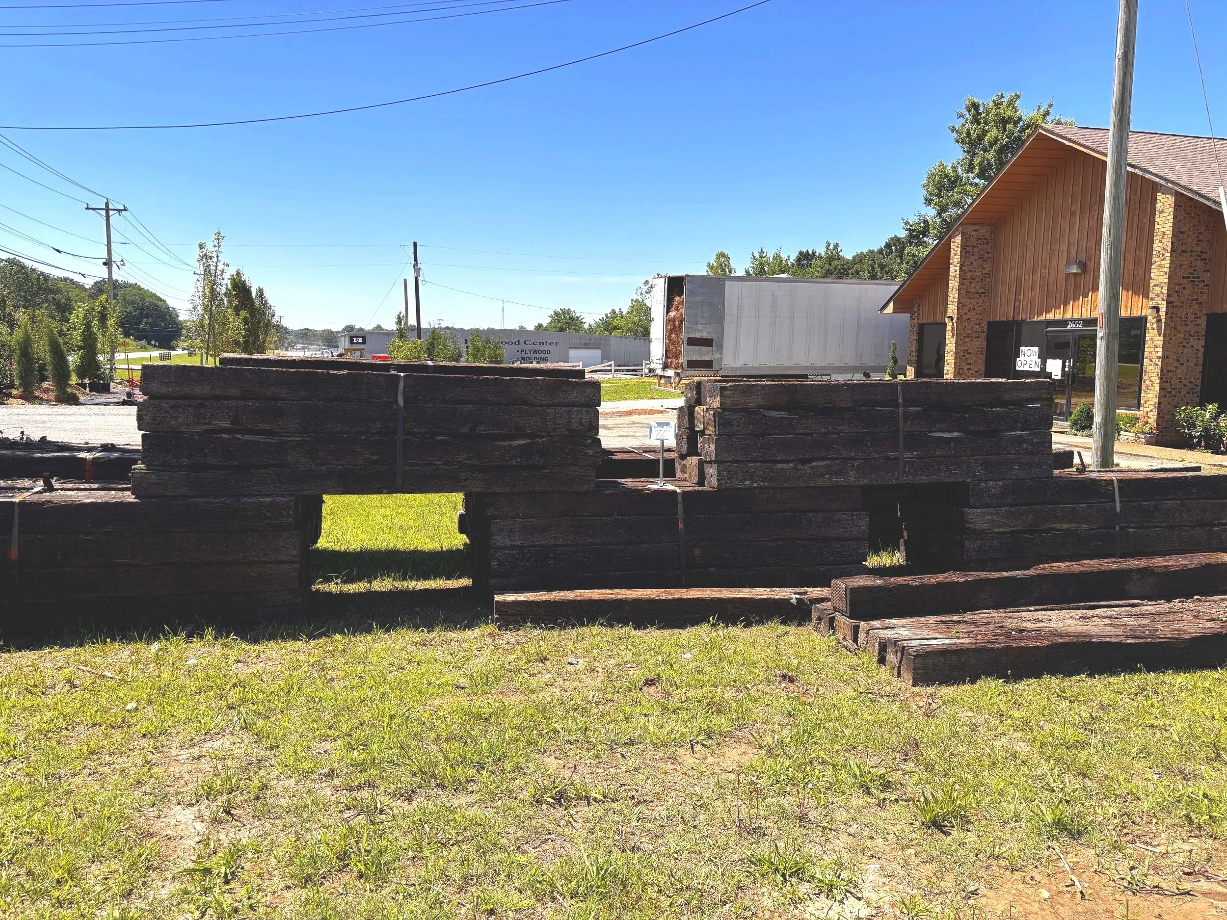 Stacked wooden planks outdoors with a brick building, trees, utility poles, and a clear blue sky in the background.