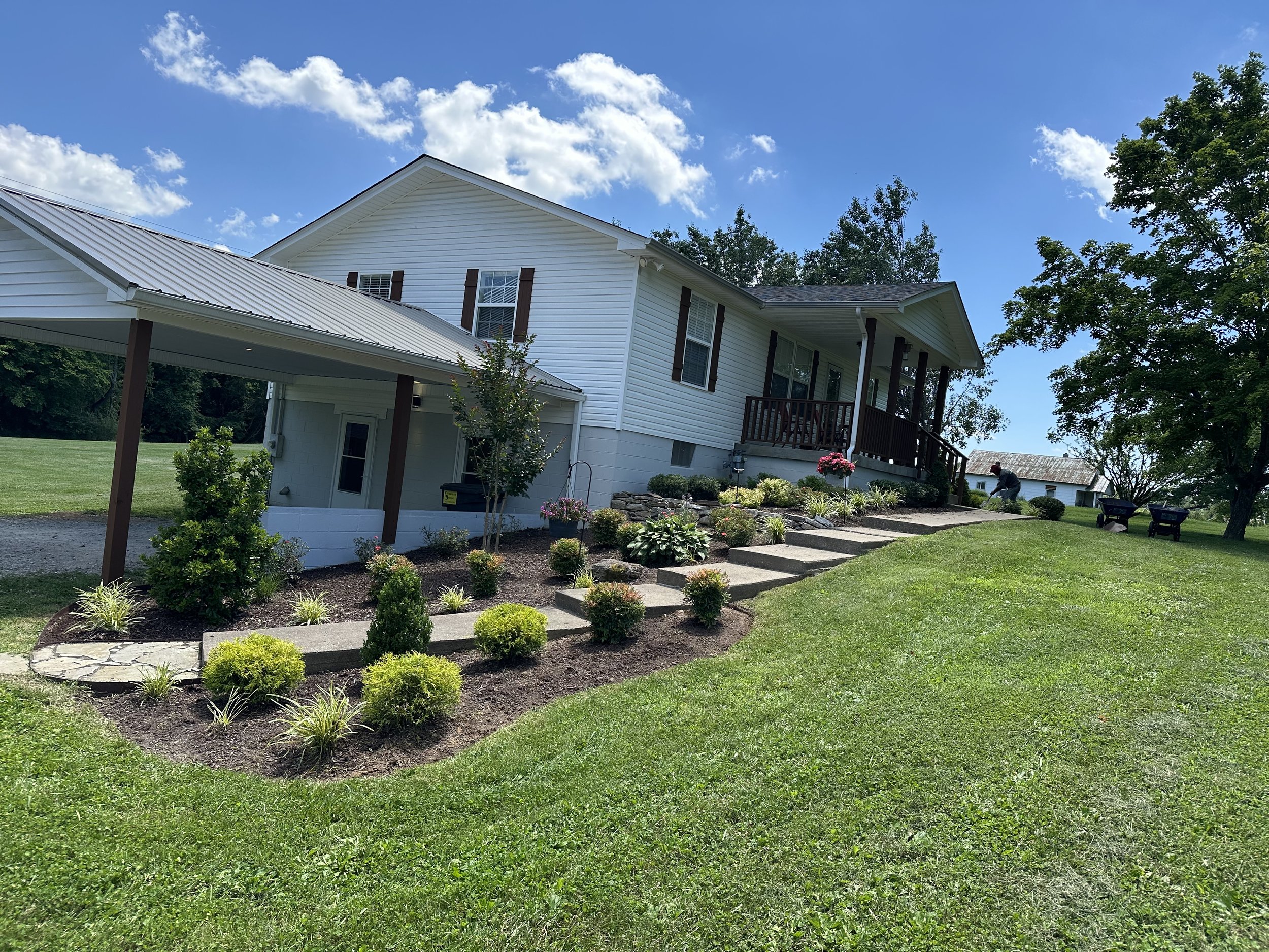 White house with a porch, set on a grassy yard with landscaping including small bushes and flowers, under a partly cloudy blue sky.