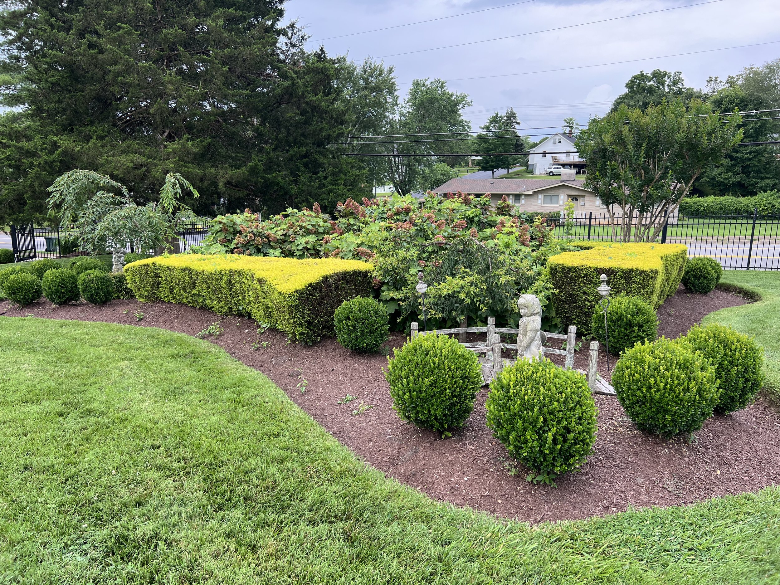 Well-maintained garden with various bushes, hedges, and trees surrounded by grassy lawn, enclosed by a black metal fence, with a small decorative stone statue of an owl near a miniature wooden fence.