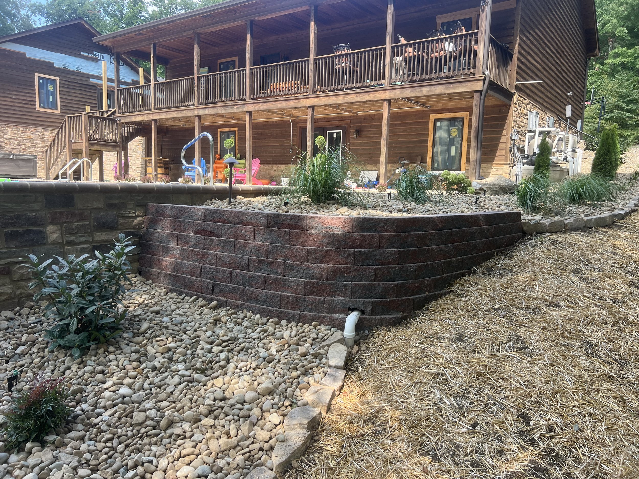 A multi-level wooden house with a stone and brick retaining wall in the foreground. The house has a large balcony with outdoor furniture, and a deck area with colorful chairs and plants. The area around the house is landscaped with rocks and plants.