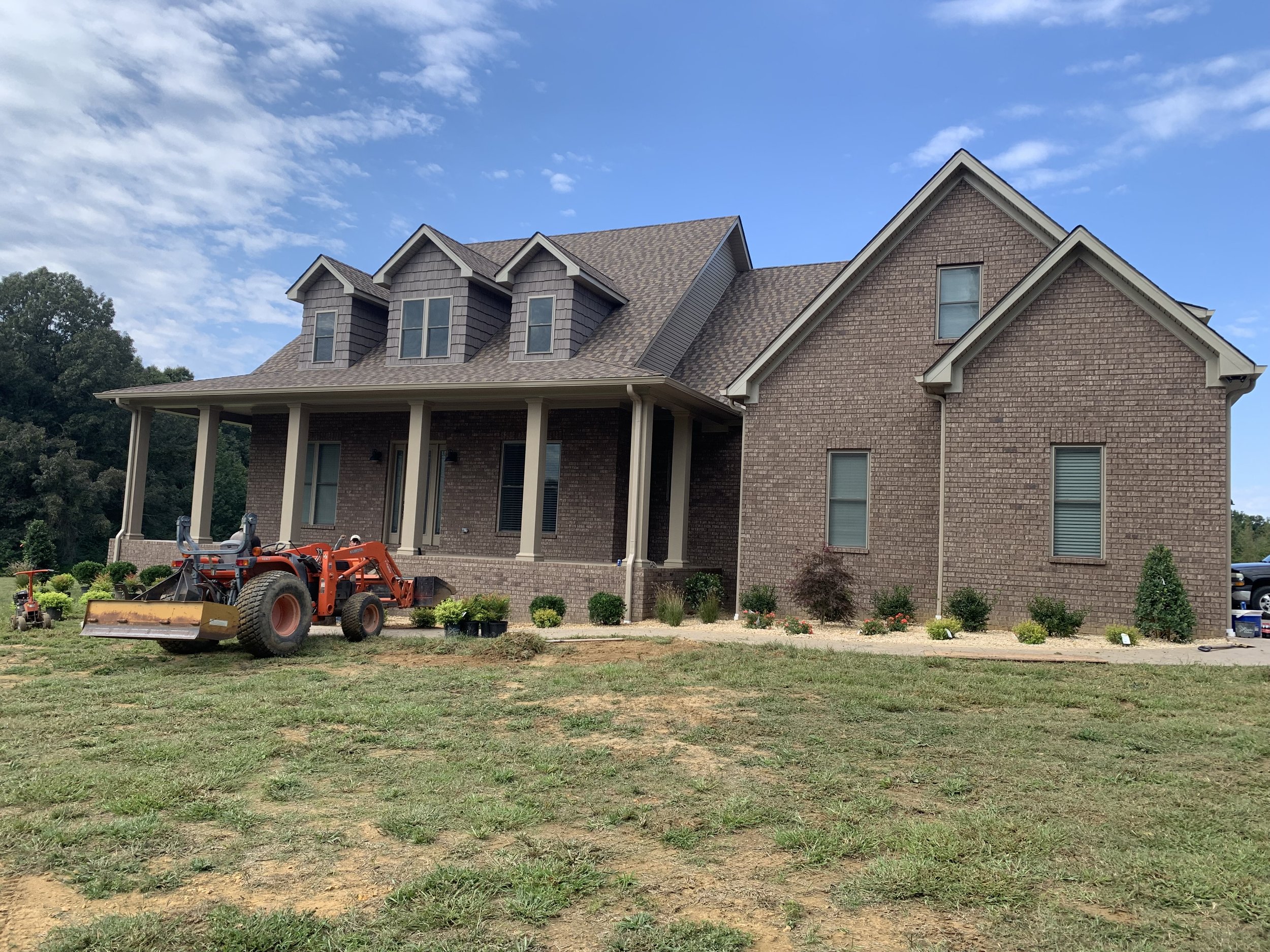 Two-story brick house with a large front porch, several windows, and a landscaped yard, with construction equipment parked in front under a blue sky.