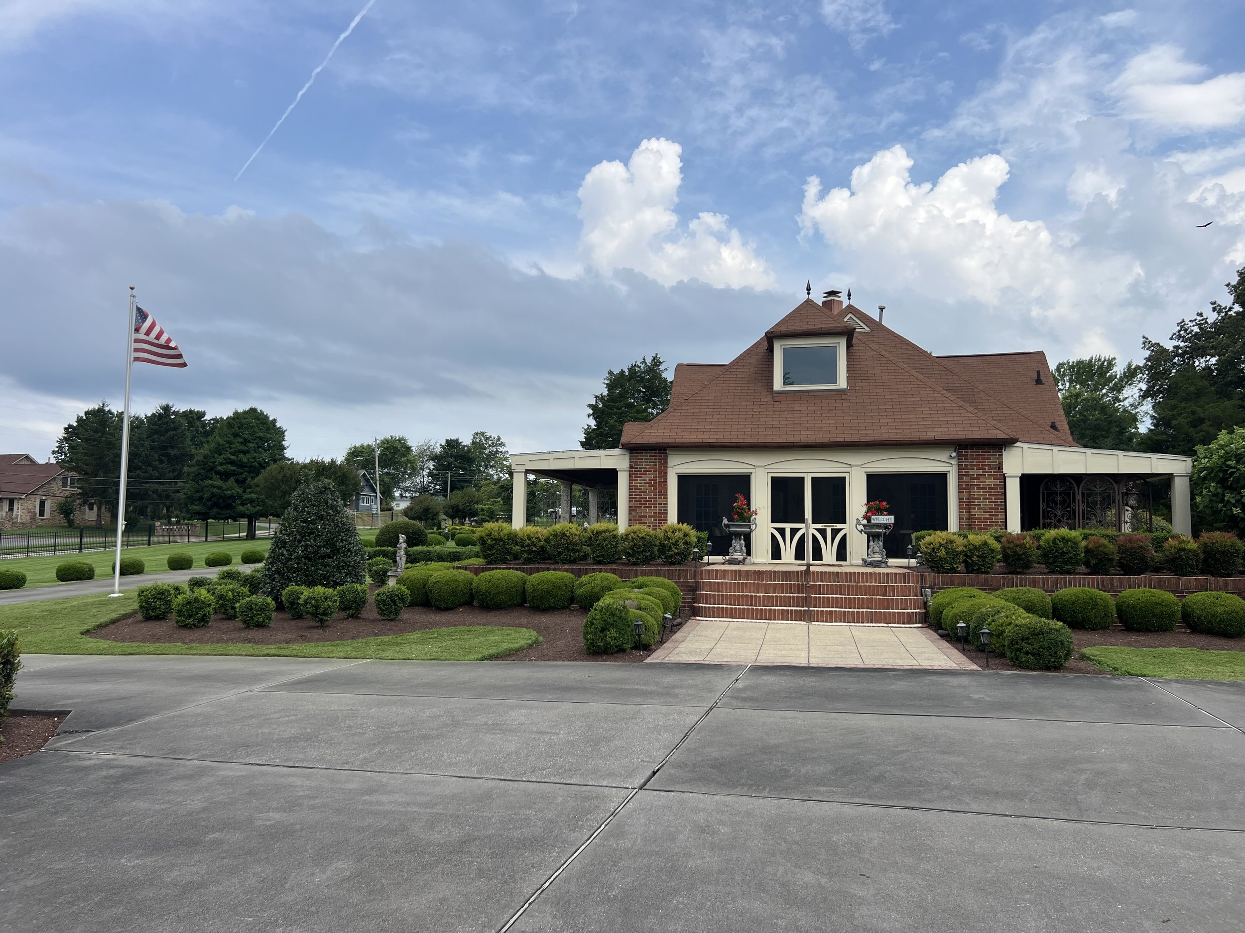 Front view of a house with a brick exterior and a brown gabled roof, surrounded by well-manicured bushes and garden decorations, with an American flag on a flagpole, under a partly cloudy sky.