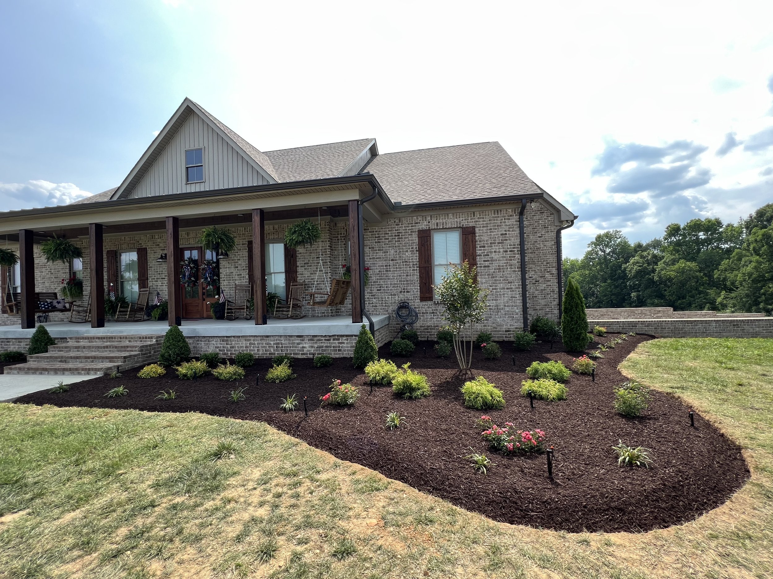 Front view of a brick house with a large front porch, featuring rocking chairs, hanging plants, and decorative wreaths. The house has a shingled roof and several windows with wooden shutters. The landscaped yard has a curved flower bed with small shr
