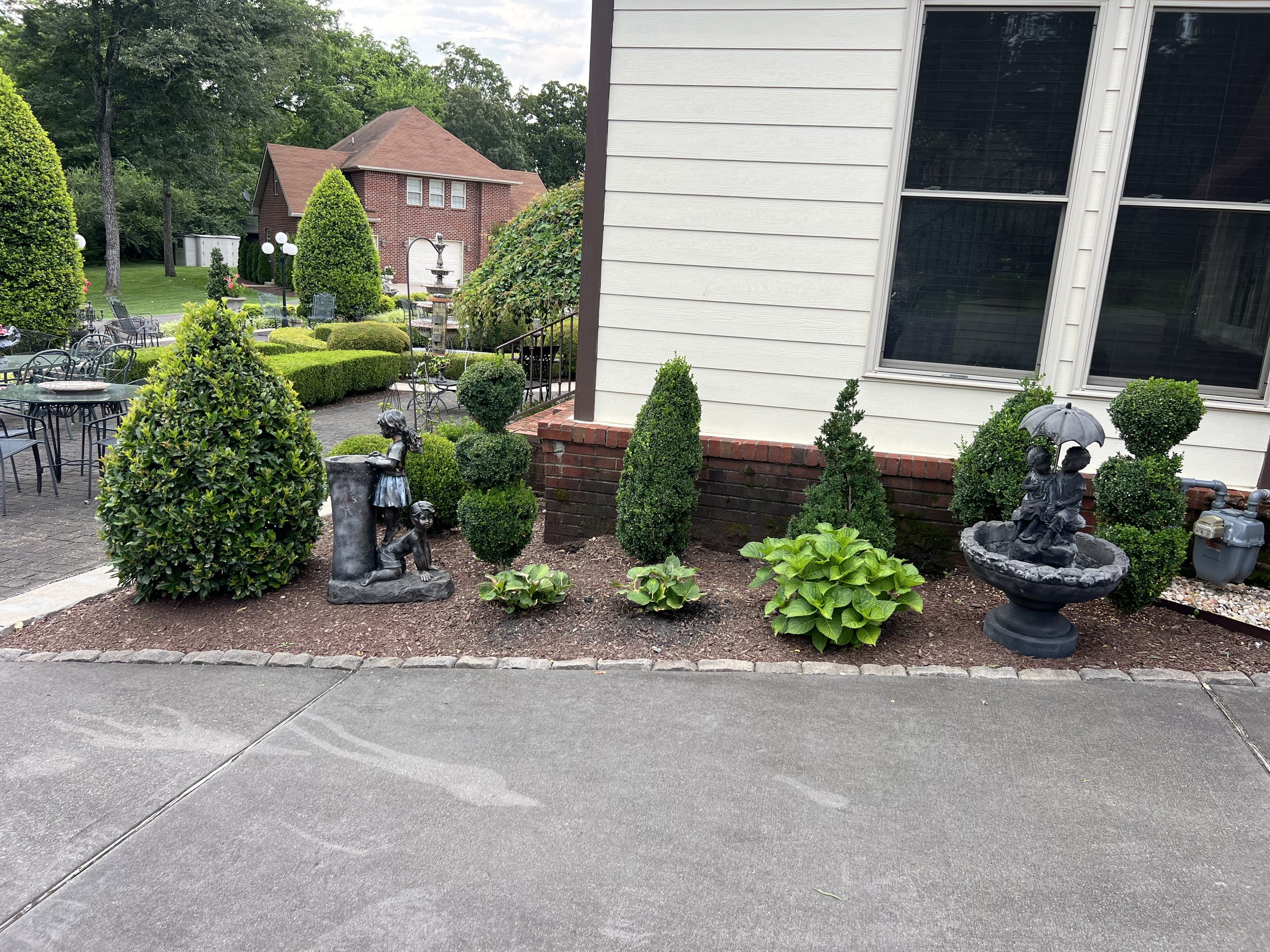 Garden bed with various green bushes, decorative statues of children, and trees in the background next to a house with white siding and large windows.