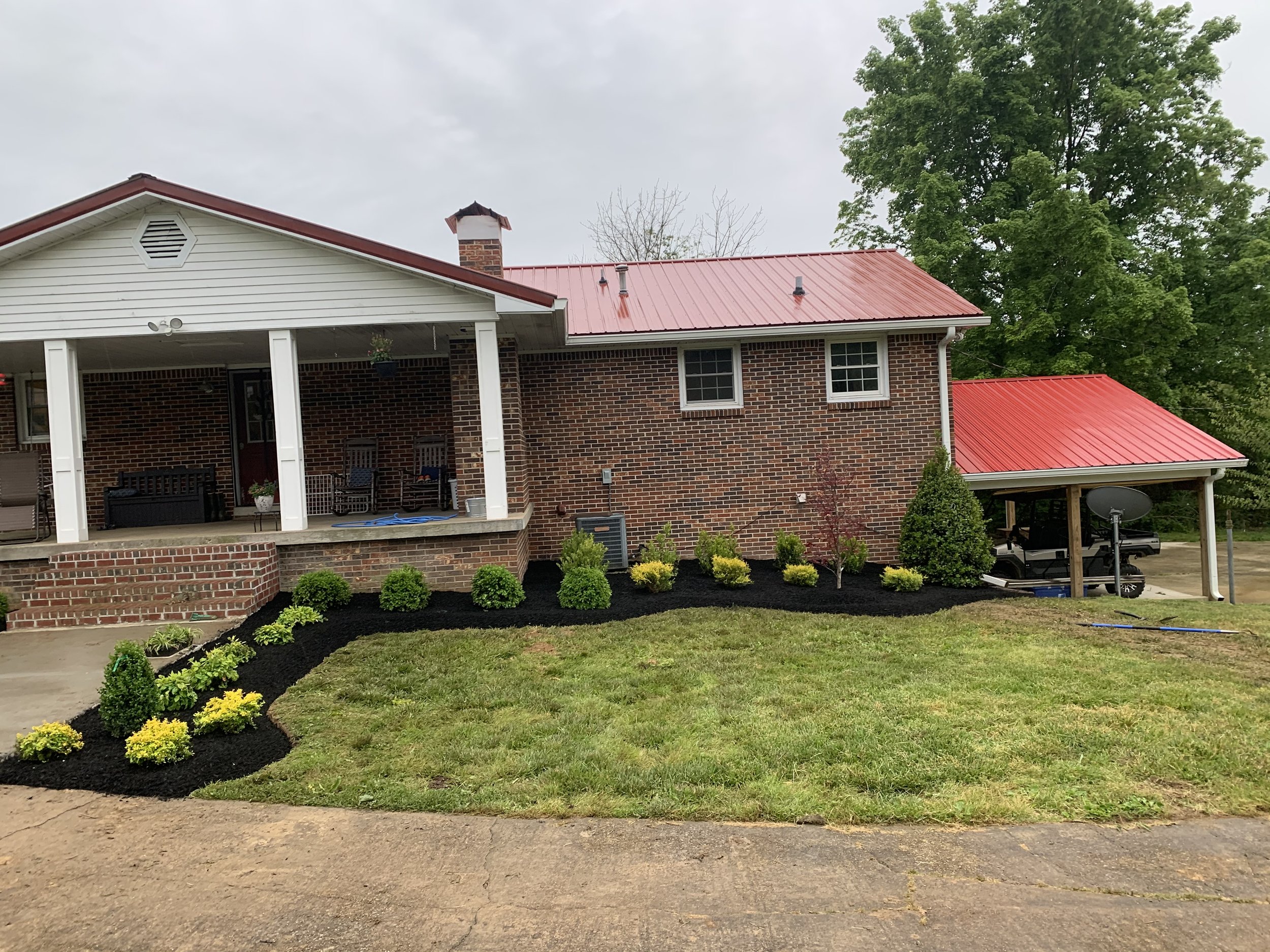 A brick house with a red metal roof, porch with chairs, and freshly landscaped garden with bushes and mulch.