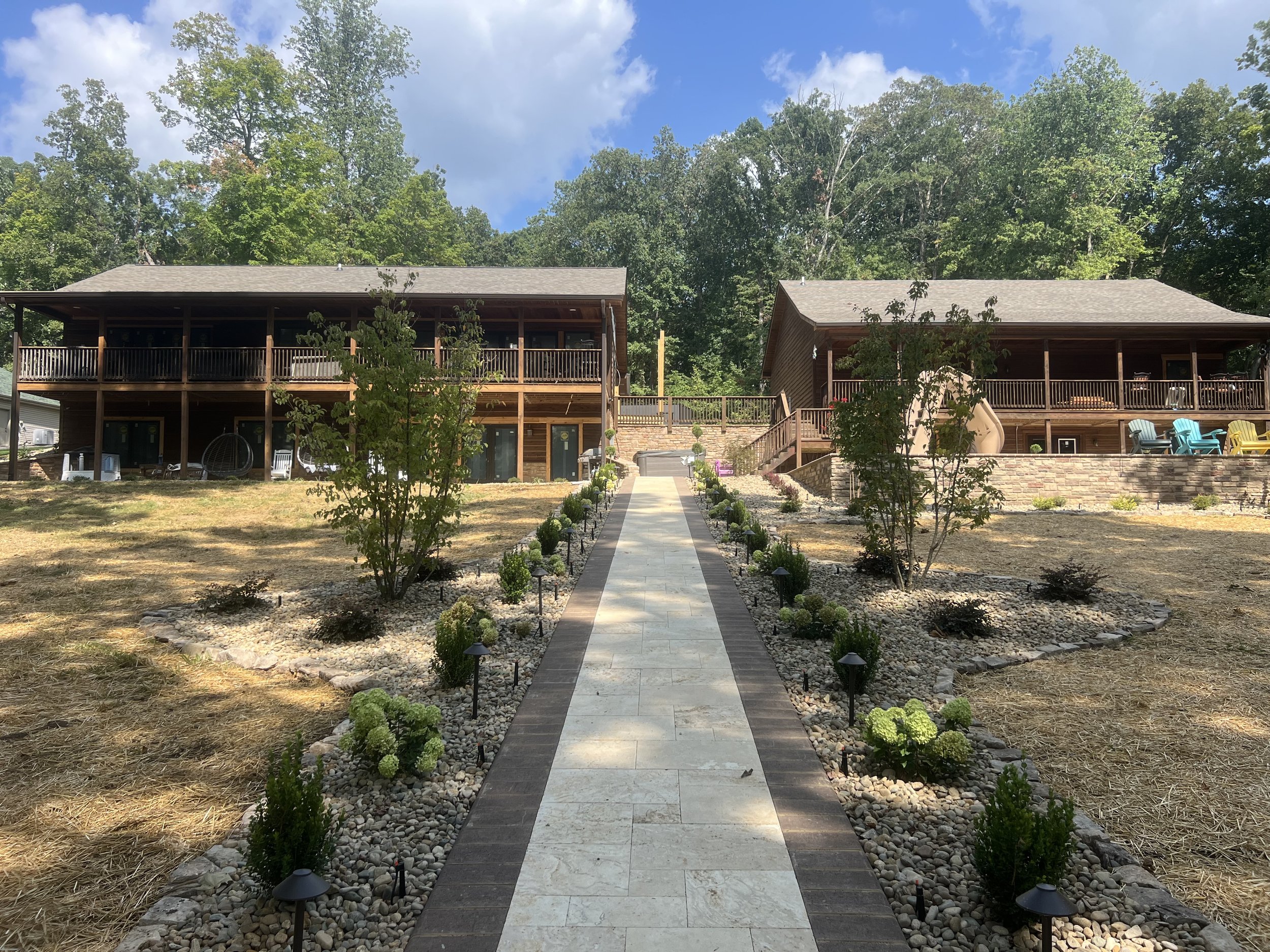 A landscaped pathway leading to two wooden houses with decks, surrounded by trees and small shrubs.