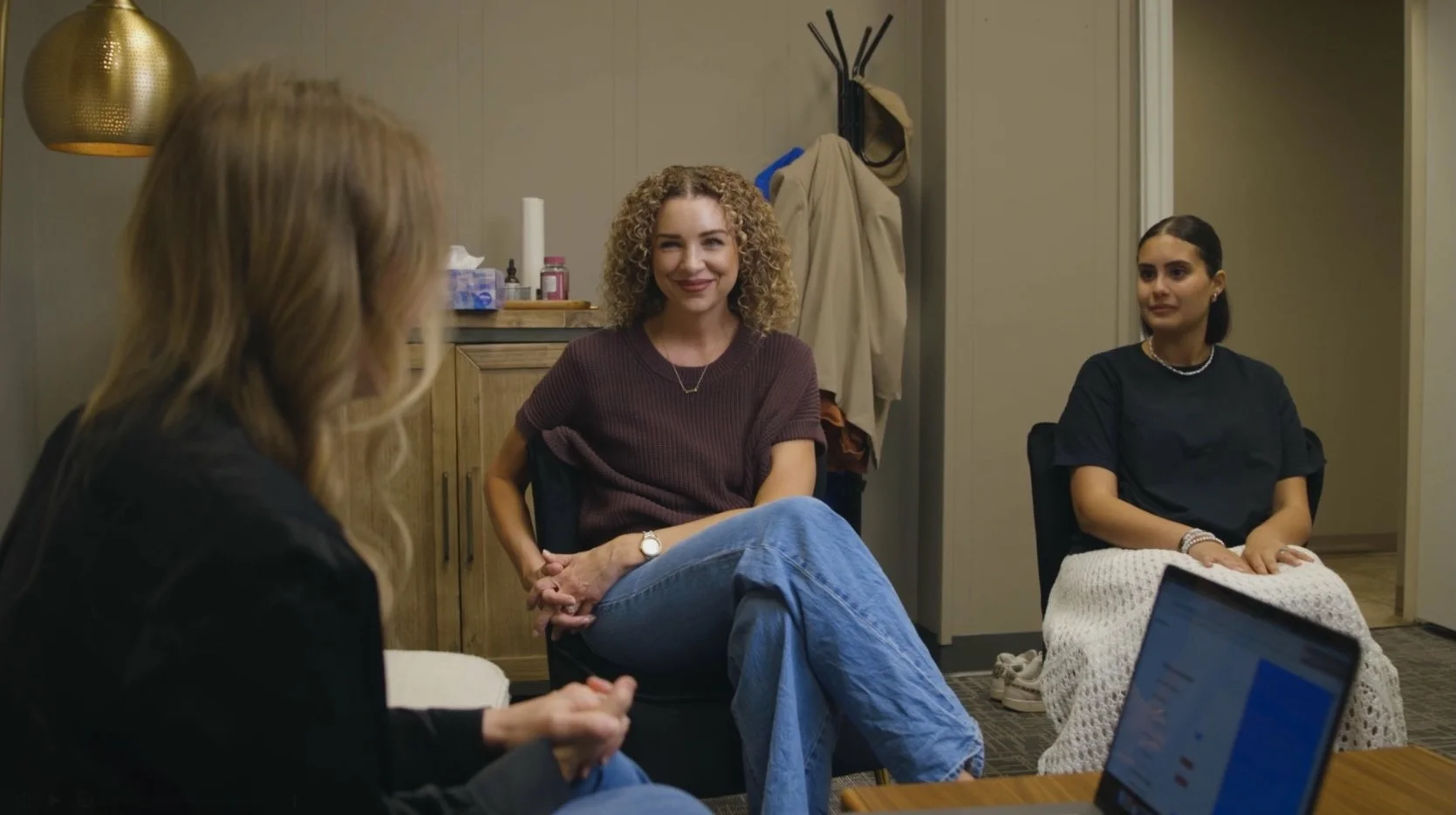 Three women seated in a circle having a conversation in an indoor setting. One woman is smiling, while the other two listen attentively. There is a laptop on a table in front of one woman.