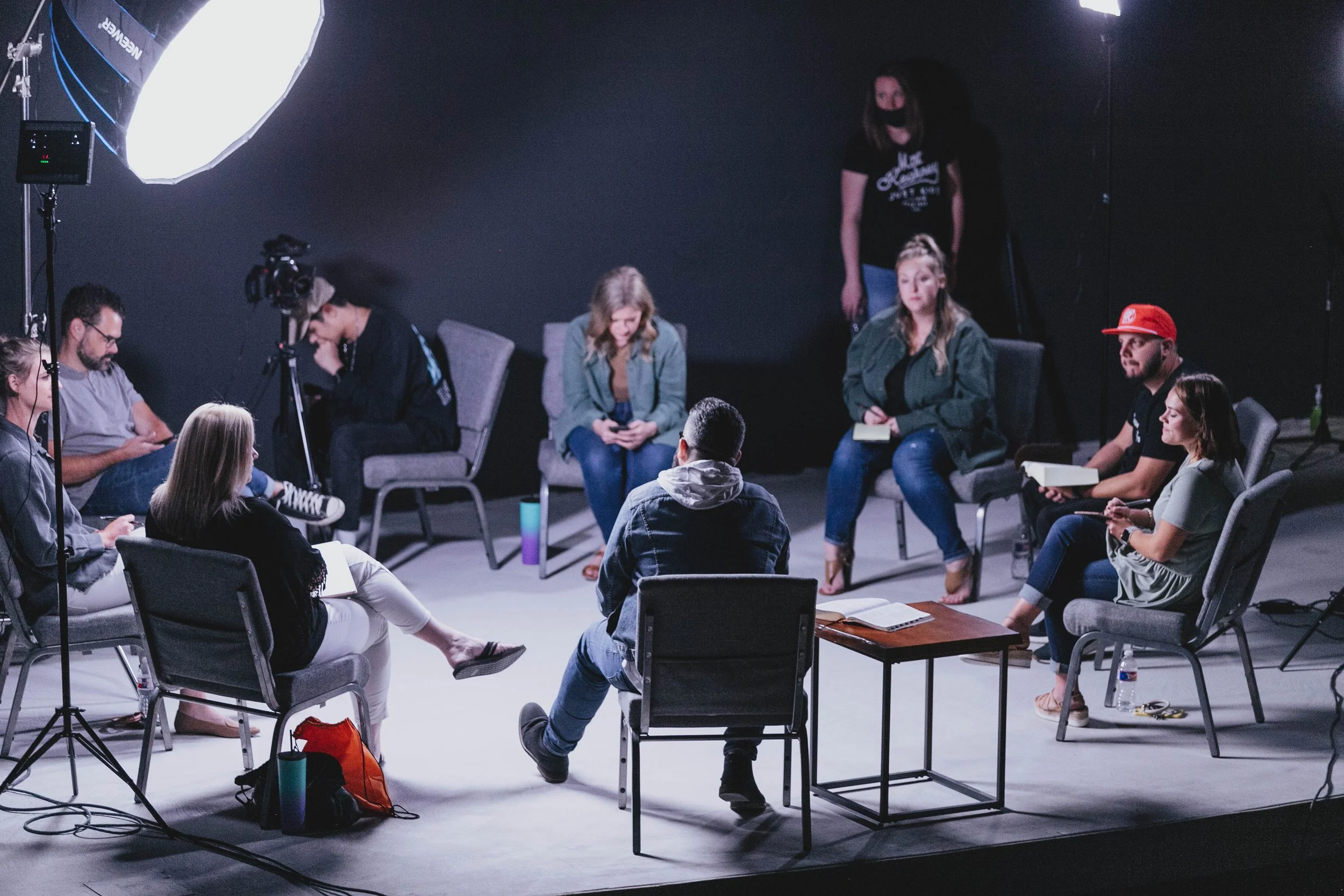 A group of nine people sit in a circle during a discussion or interview, with two large studio lights overhead and a camera recording. The setting appears to be a studio with a black wall background.