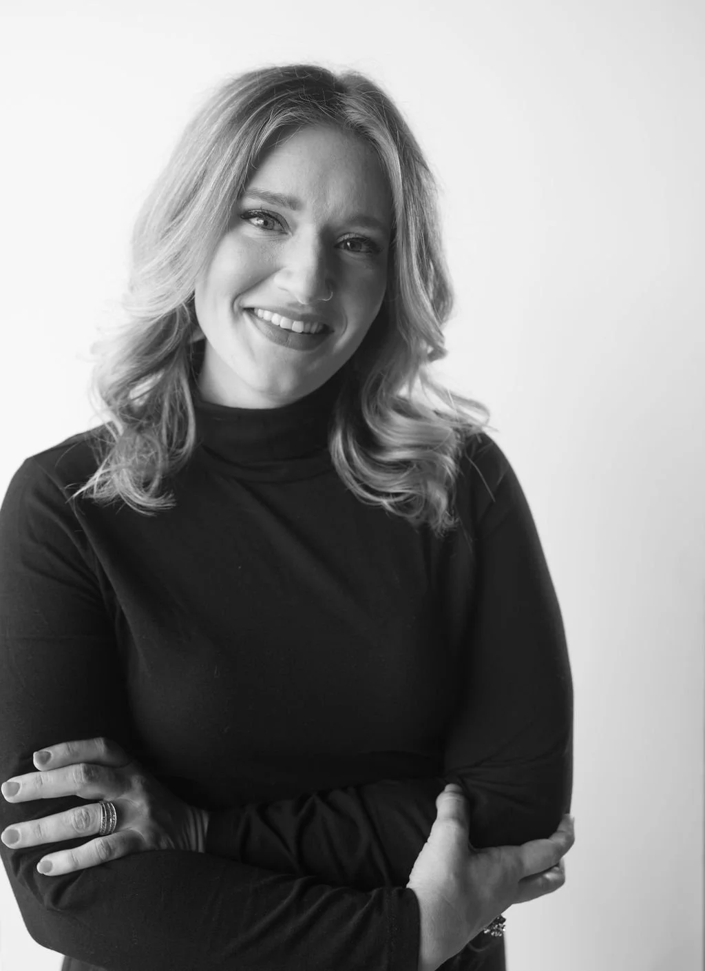 Black and white portrait of a smiling woman with wavy hair wearing a black turtleneck, standing against a plain white background.