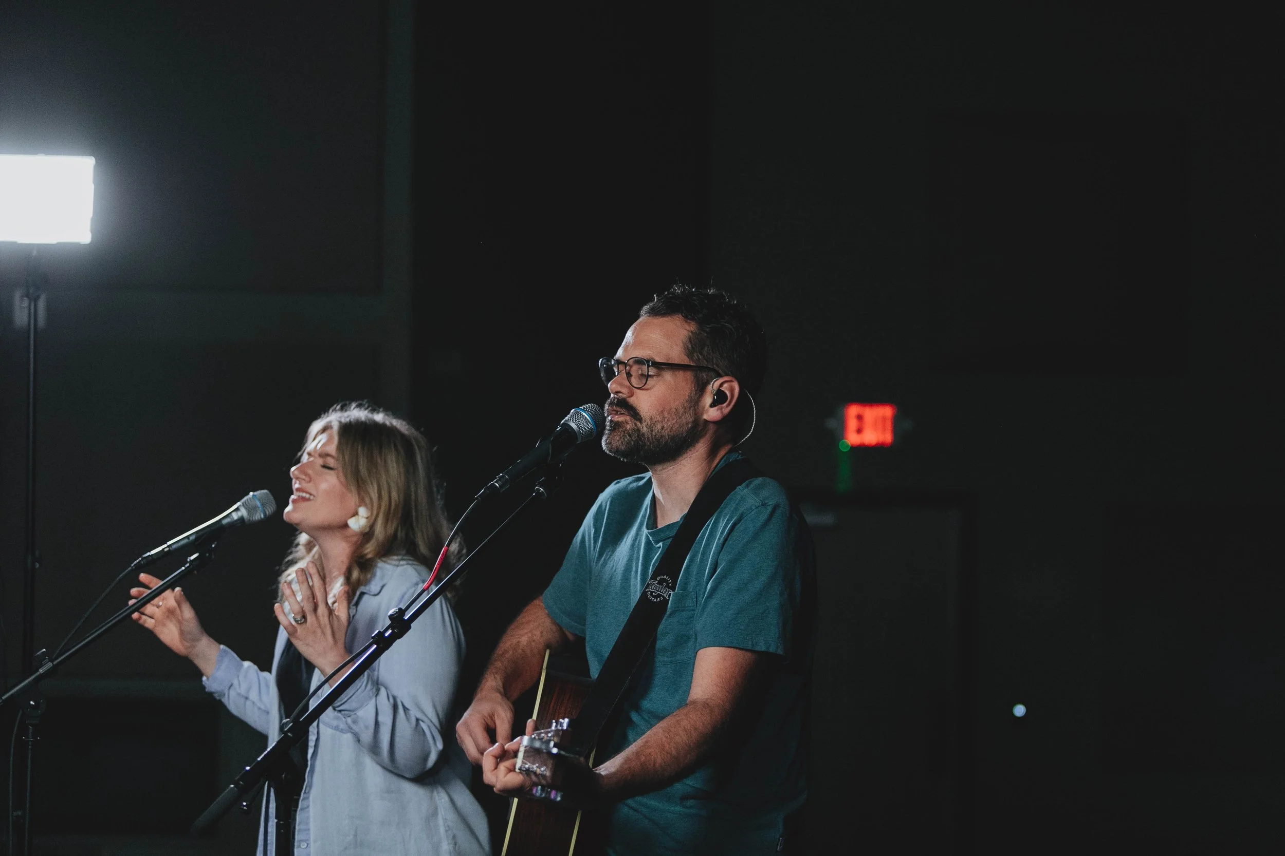 Man and woman performing music on stage, woman singing into microphone, man playing guitar with eyes closed, dark background with exit sign