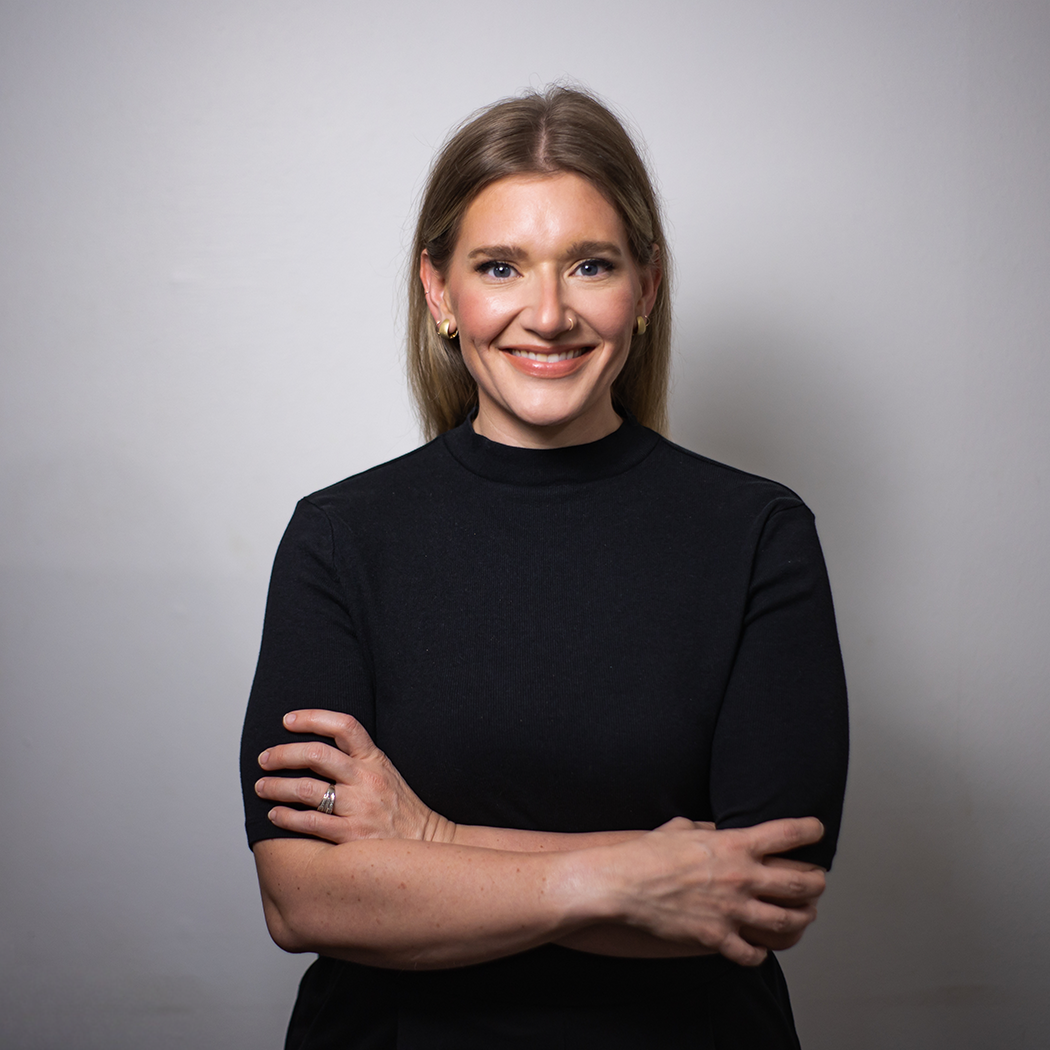 A woman with long blonde hair smiling, dressed in a black top with her arms crossed, standing against a plain gray background.