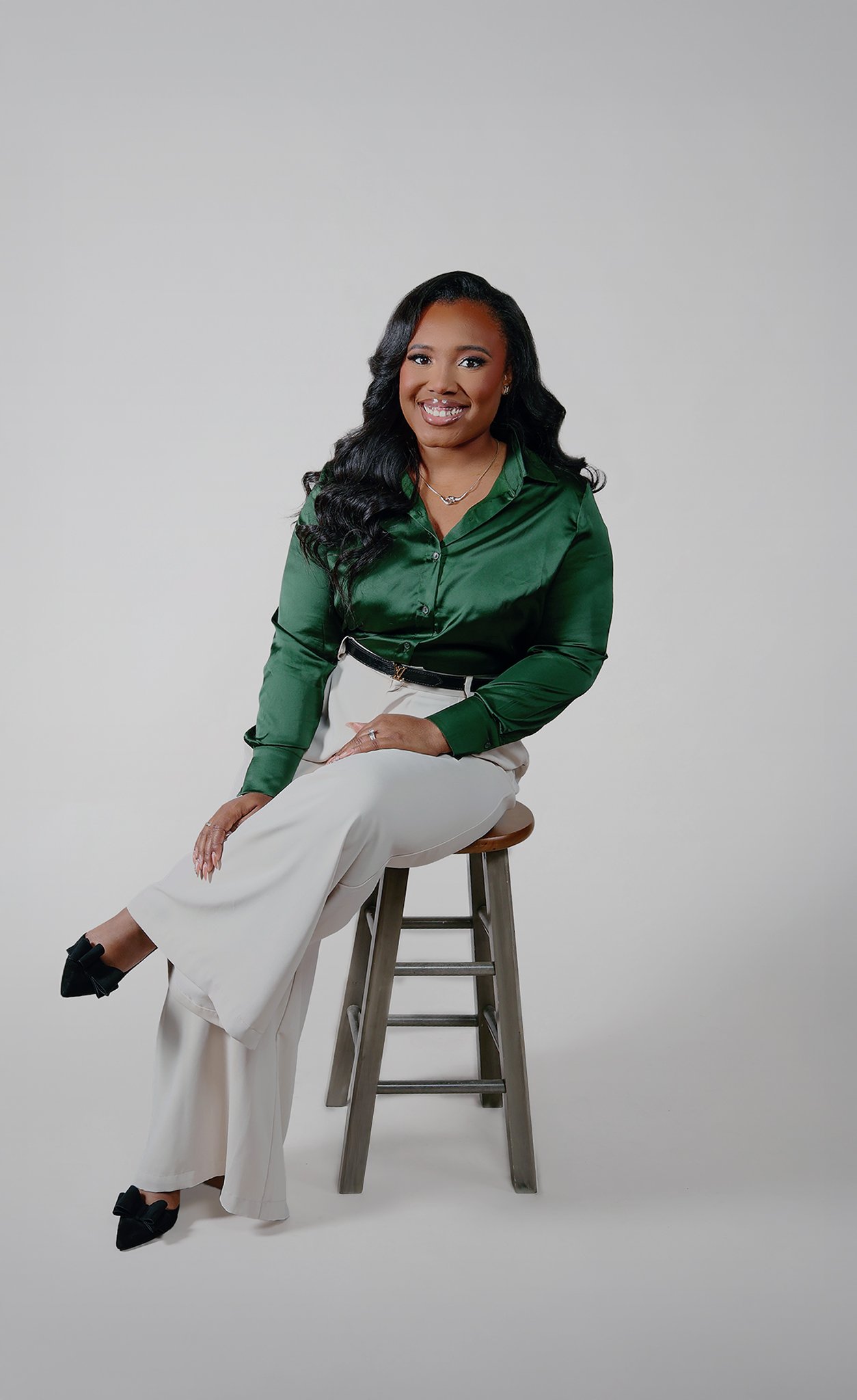A woman with dark, wavy hair sits on a wooden stool, wearing a green satin blouse, white wide-leg pants, and black heels with bows, smiling at the camera against a plain white background.