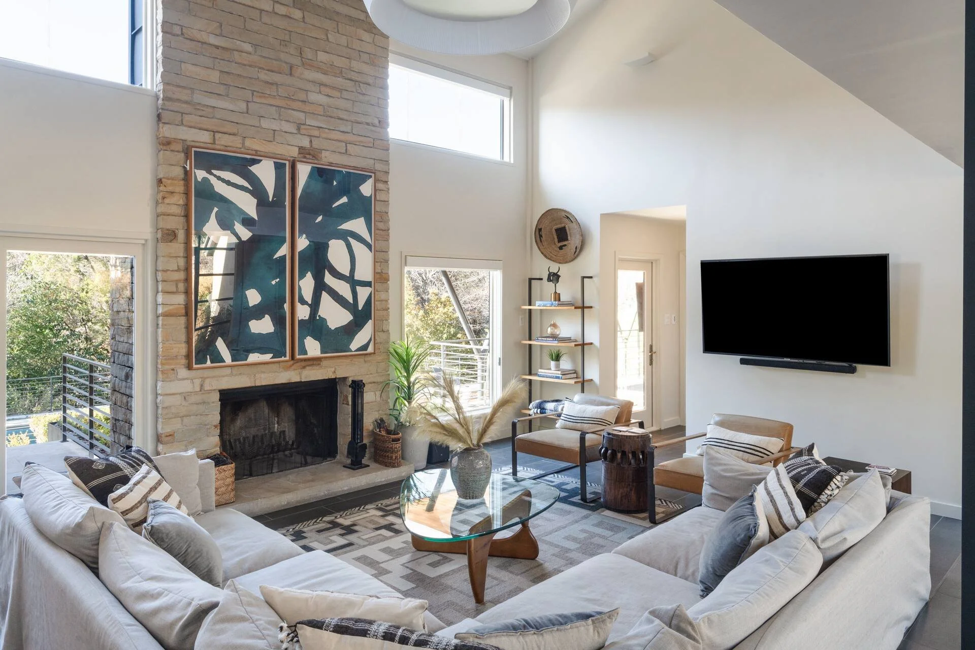 Living room with a stone fireplace, a large abstract art piece above it, beige couches with patterned pillows, a glass coffee table, an armchair, a wooden side table, a TV on the wall, and large windows letting in natural light.
