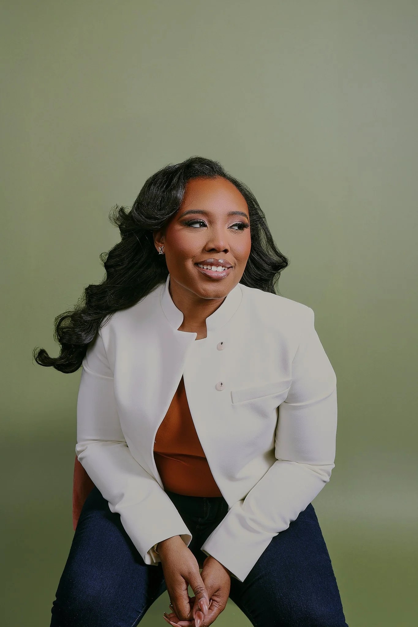 A woman with dark, wavy hair wearing a white blazer, a brown top, and dark jeans sitting against a light green background, smiling slightly and looking to the side.