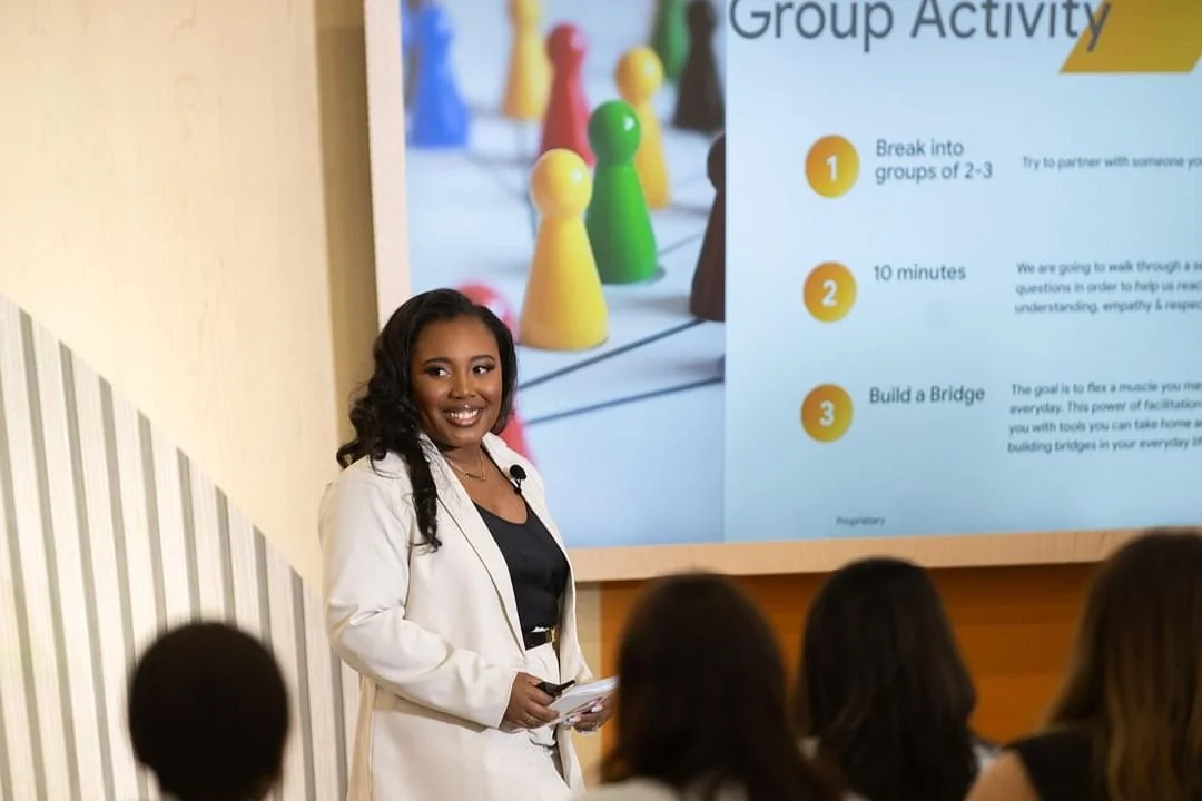 A woman with dark curly hair, wearing a white blazer, standing and smiling in front of a screen during a group activity presentation. The screen displays colorful game pieces and instructions for a team-building exercise.