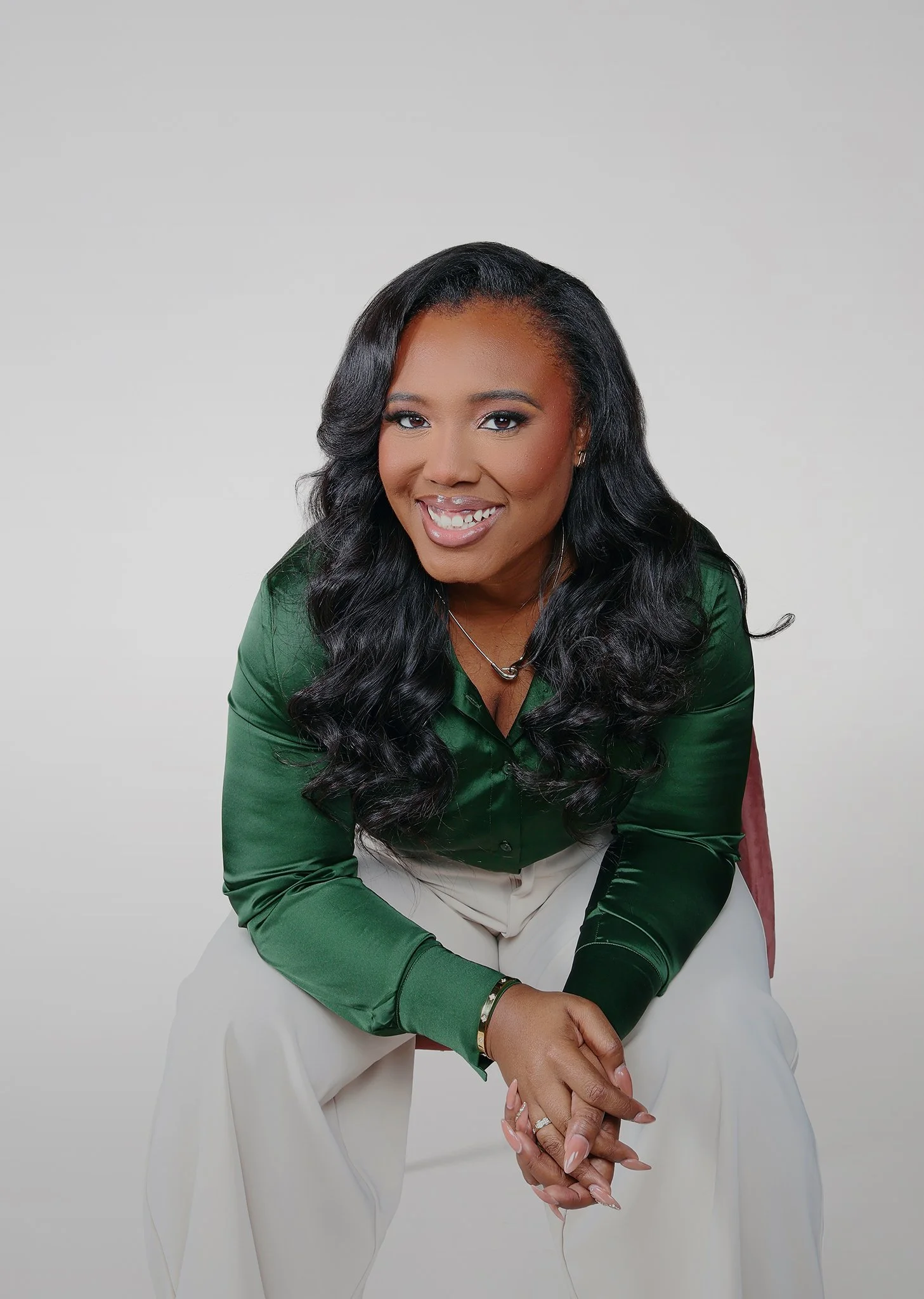 A woman with long, wavy black hair smiling, wearing a green satin blouse and white pants, sitting against a plain gray background.