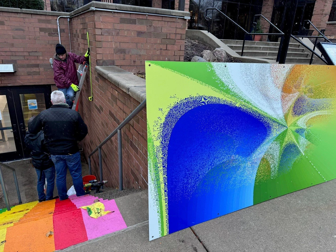 Two men working on construction or installation outside a building. One man is on a ladder, and they are working near a colorful painted wall or panel with a rainbow gradient. The blue, green, and orange colors dominate the artwork.