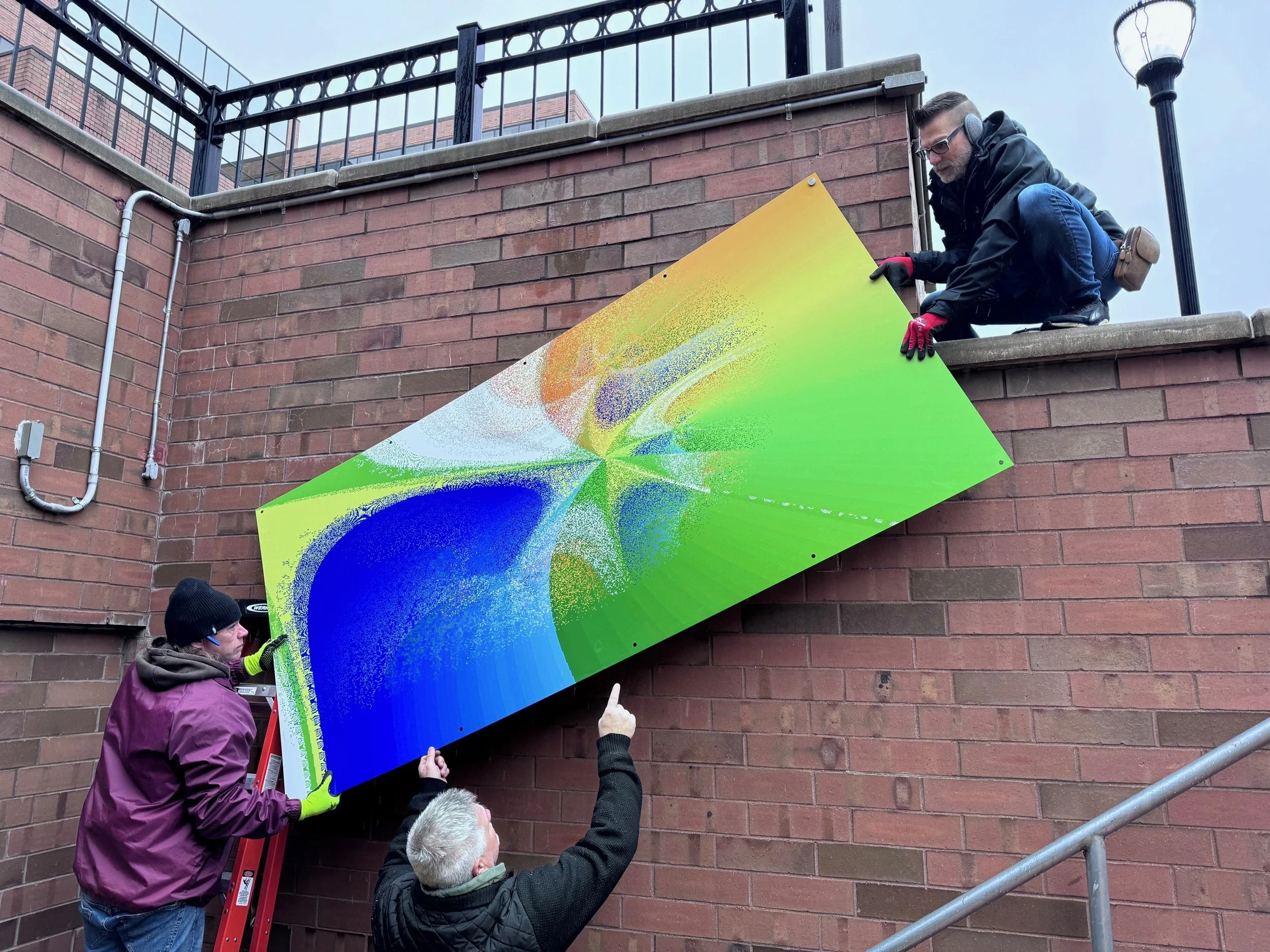 Three men are installing a colorful abstract artwork on a brick wall outdoors. One man is on a ladder, while the other two are on the ground helping.