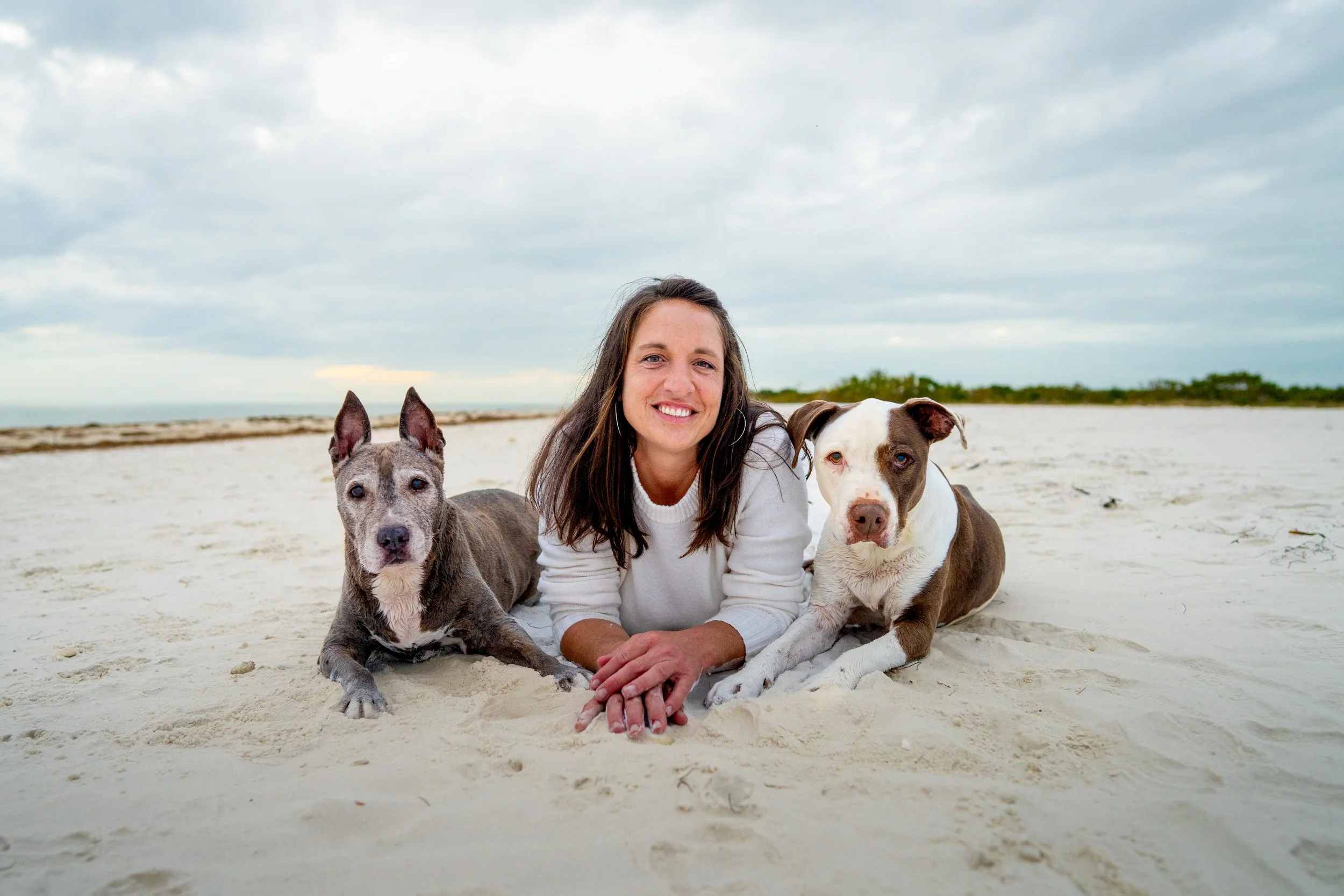 A woman with long brown hair and a white sweater smiling while lying on the sand at the beach with two dogs, a gray and white one and a brown and white one, under cloudy skies. Pictures with senior dogs