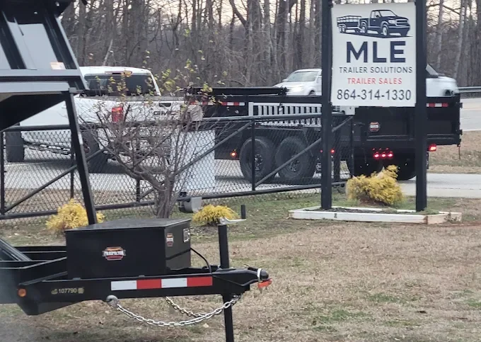 Outdoor scene showing a trailer, a chain link fence, a sign for MLE Trailer Solutions, and several vehicles on a road in the background.